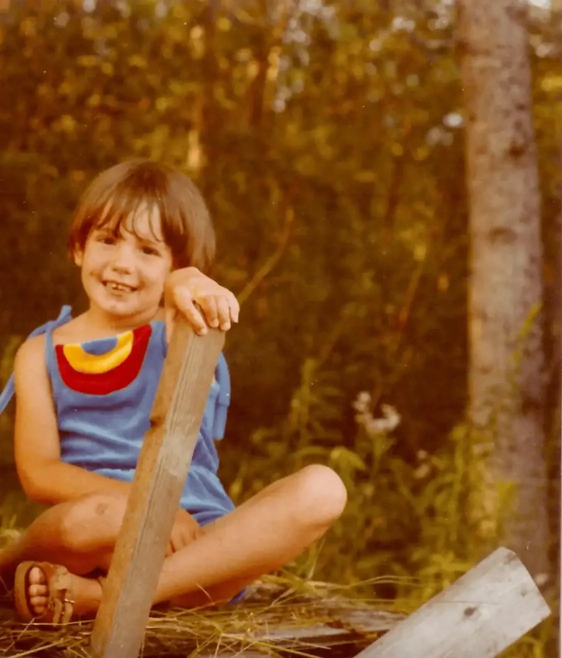 4-5 year old child in a forest siting on wood wearing a velvet outfit smiling while holding onto a piece of 2x4.