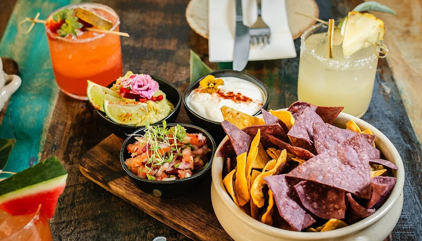 A platter of Mexican snacks including a bowl of multicolored tortilla chips, two small bowls of salsa and sour cream, and two cocktails with fruit garnishes on a rustic wooden table.