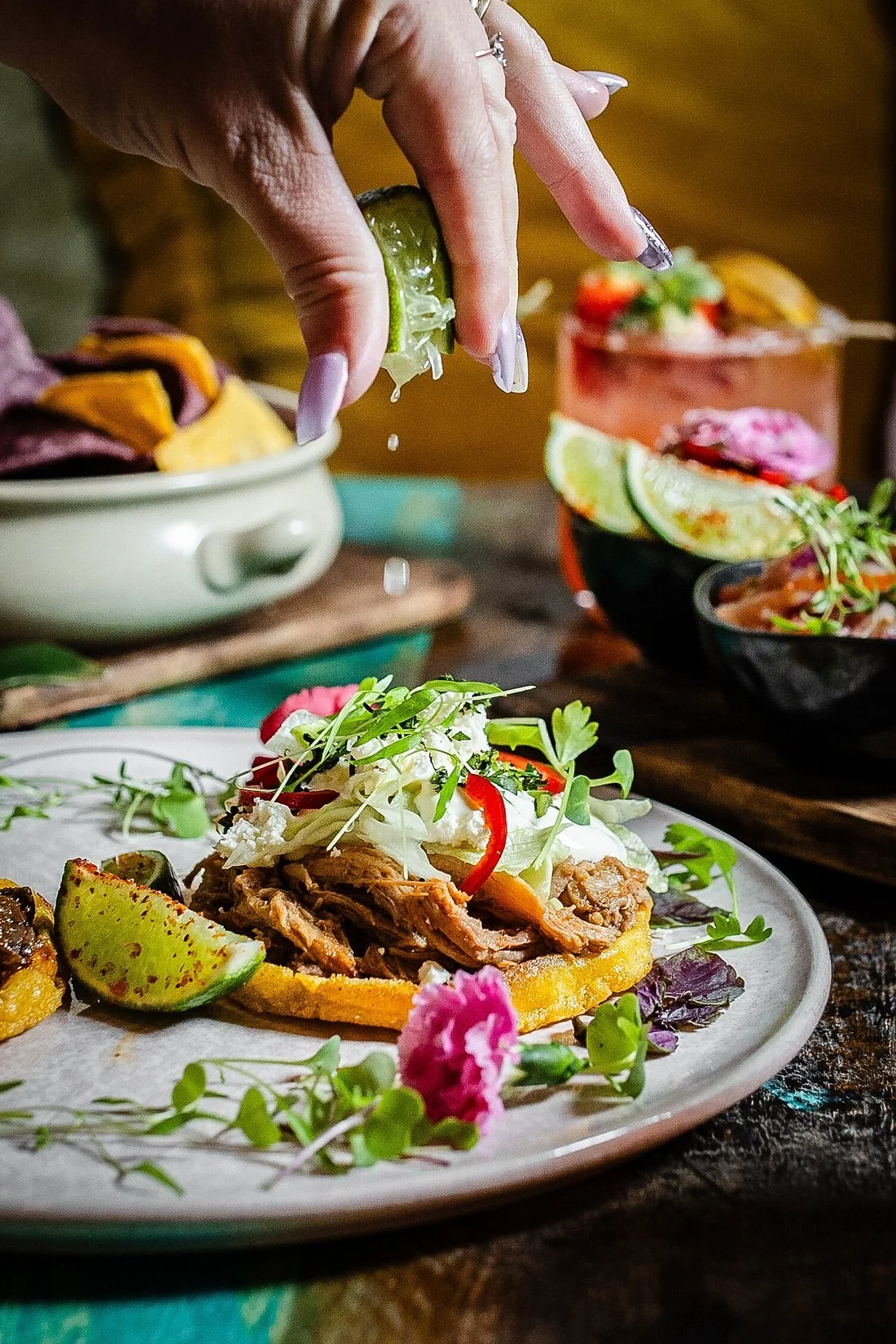 A hand squeezing a lime over a plate of food, which includes a tostada with meat, sour cream, sliced red chili, microgreens, and a wedge of lime. In the background, there are colorful drinks and bowls of garnishes.