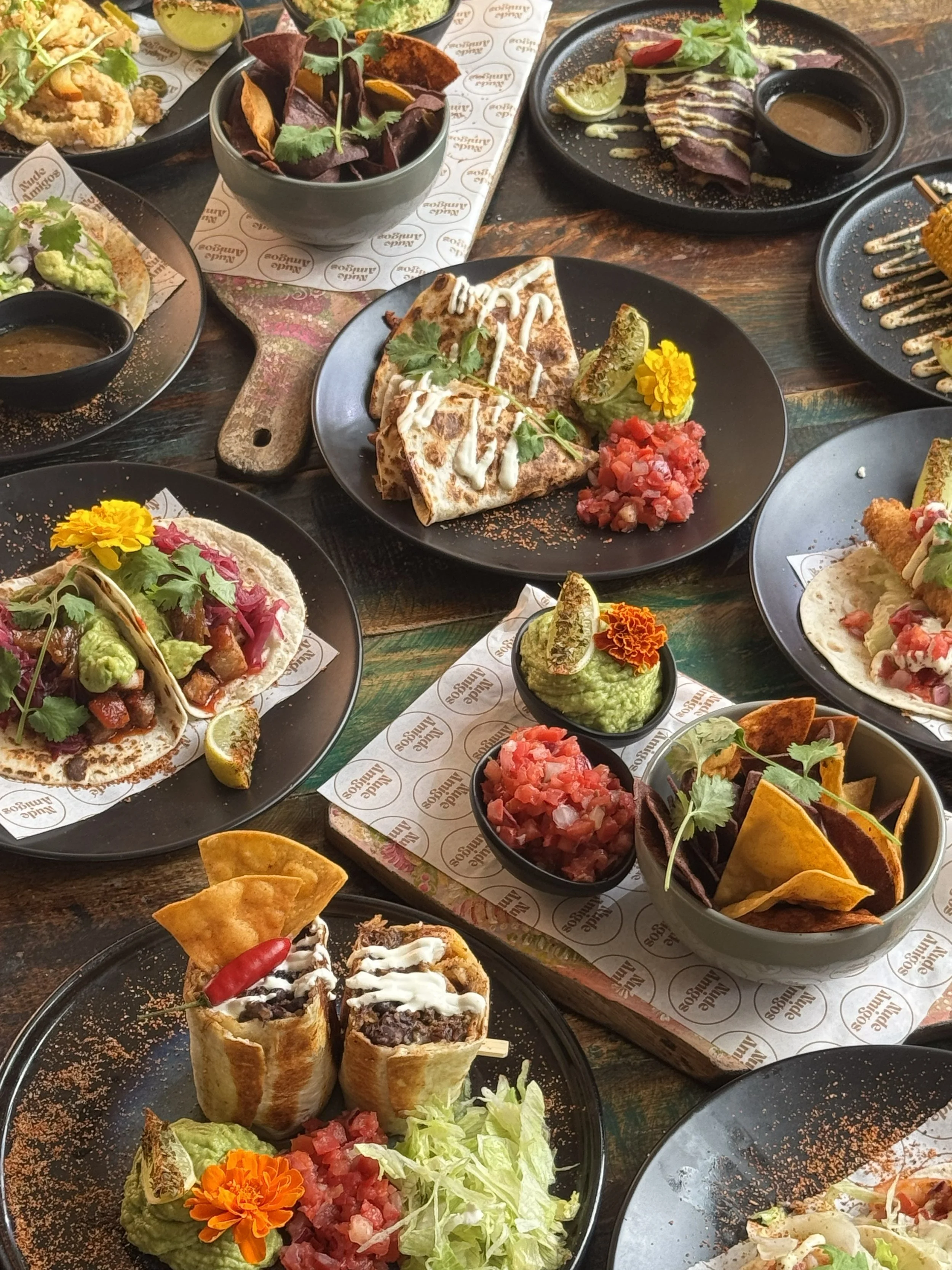 A variety of Mexican dishes including tacos, quesadillas, and a burrito, along with bowls of chips, salsa, and guacamole on a dark wooden table.