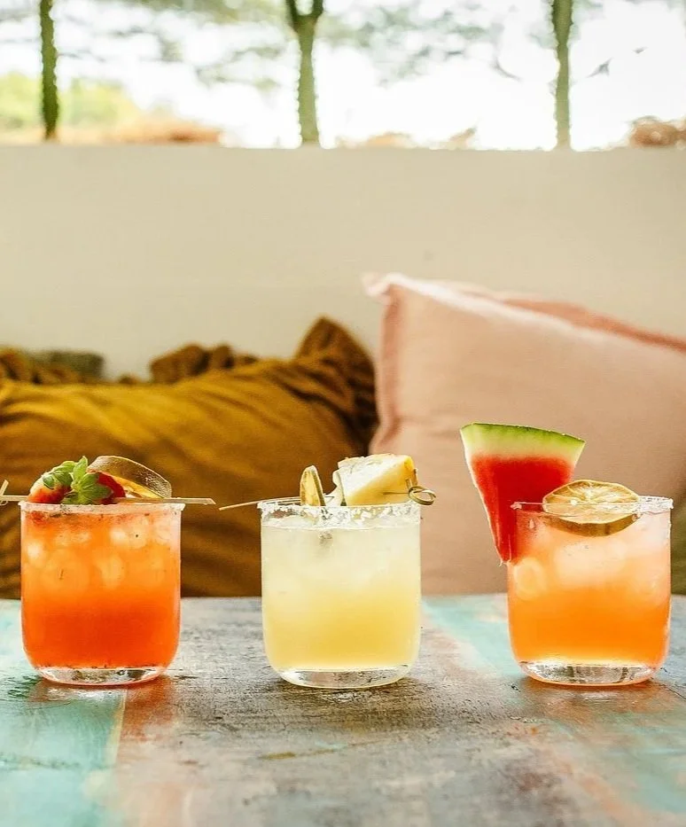 Three colorful cocktails with fruit garnishes on a table, with a blurred background of pillows and windows.