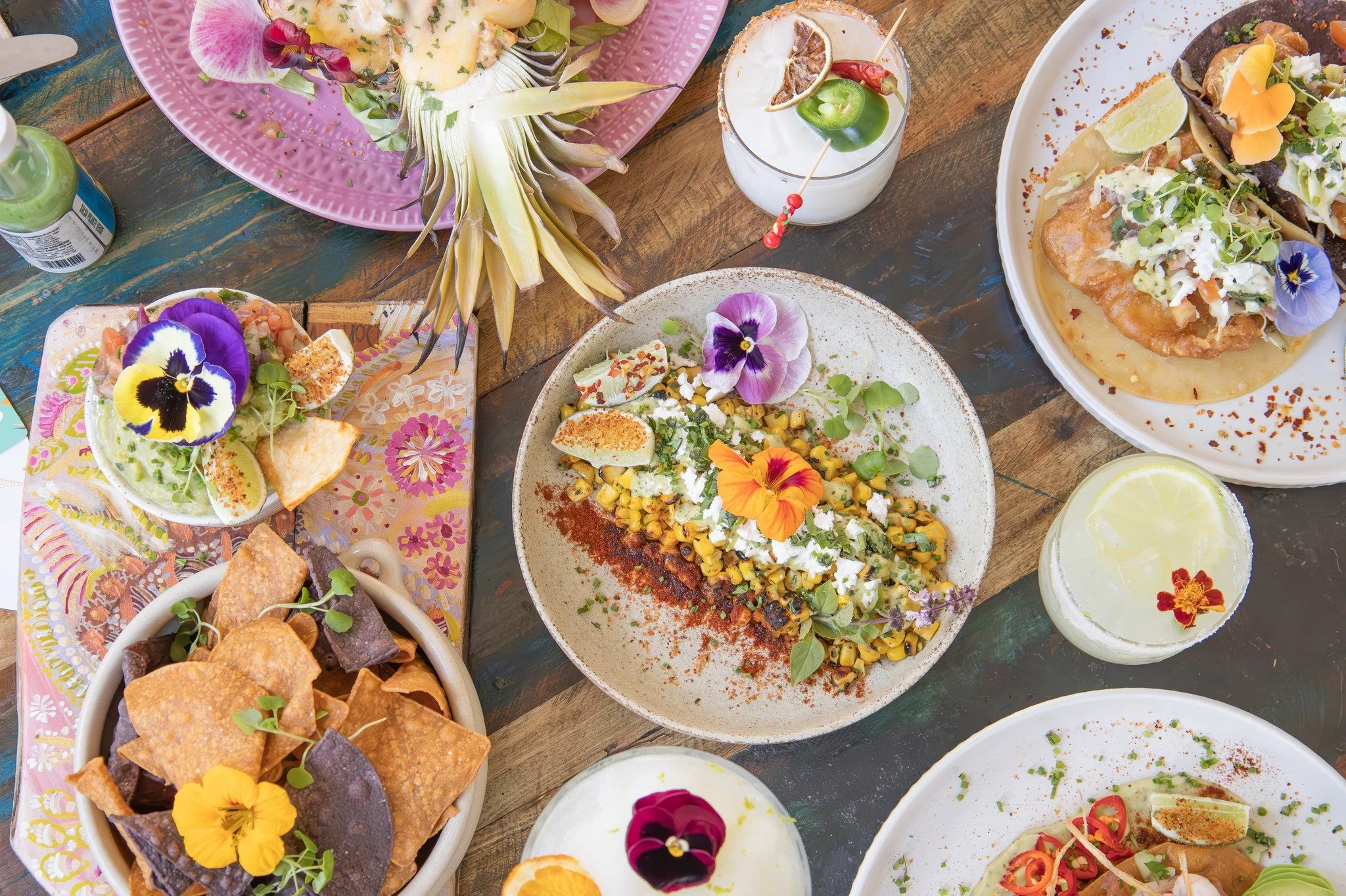 Assorted tacos with garnishes, tortilla chips, a pineapple, and beverages on a wooden table, decorated with edible flowers.