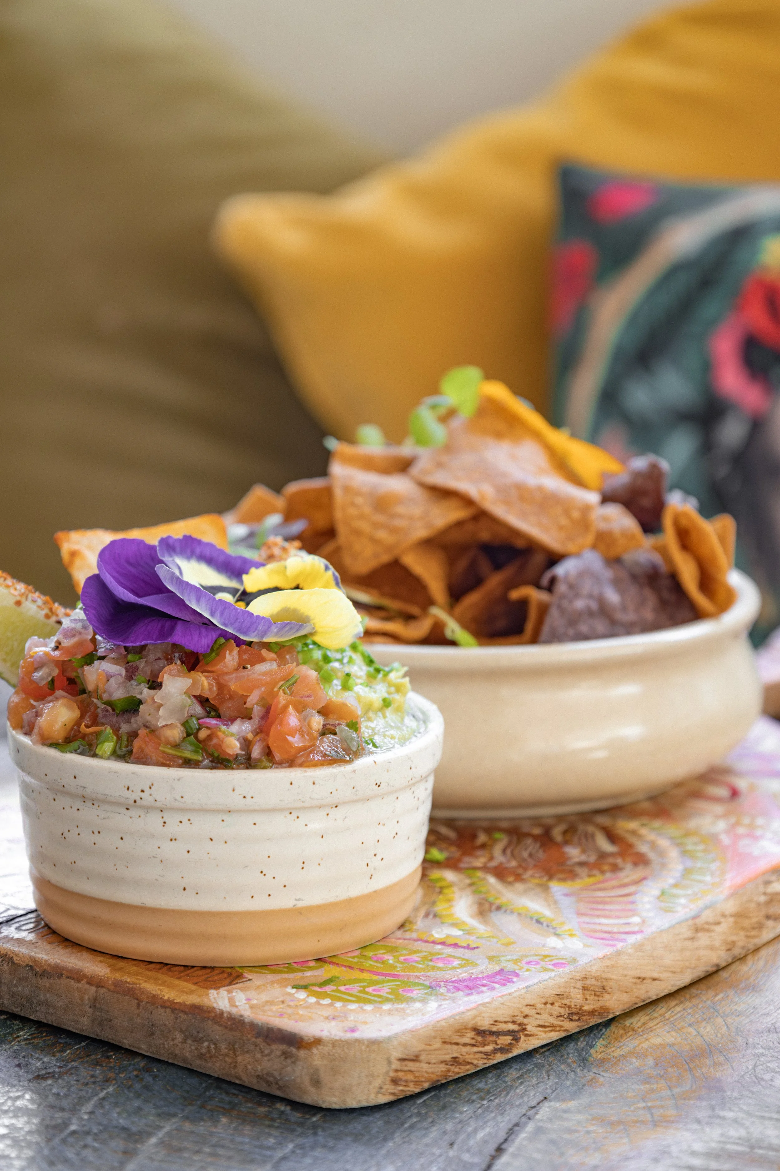 A small bowl of pico de gallo topped with edible flowers and a larger bowl of tortilla chips with microgreens, placed on a wooden serving board.