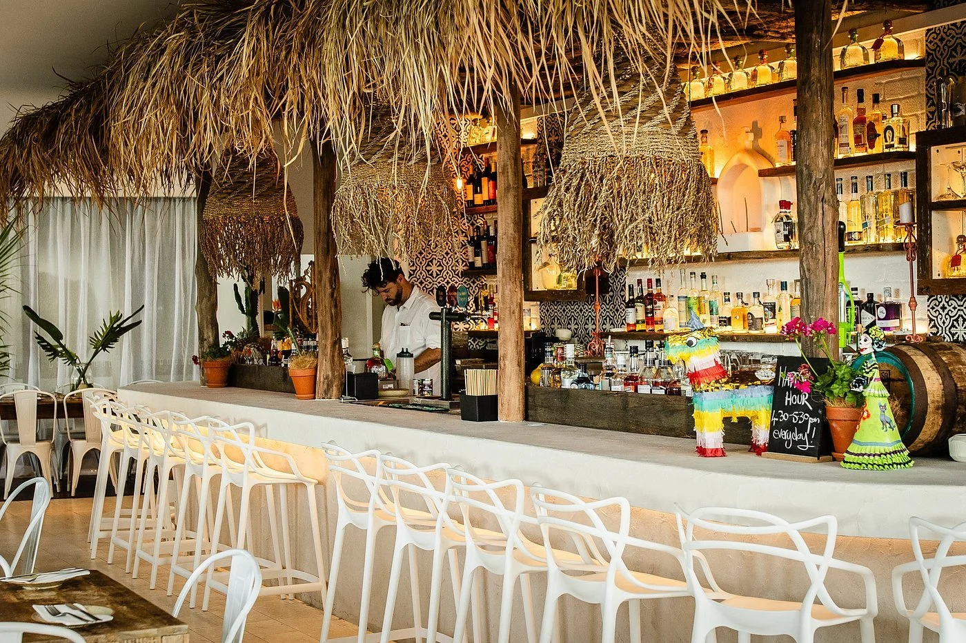 Interior of a tropical-themed bar restaurant with a thatched roof ceiling, a bartender preparing drinks, and decorative piñatas on the counter. The bar shelves are stocked with various bottles of alcohol.