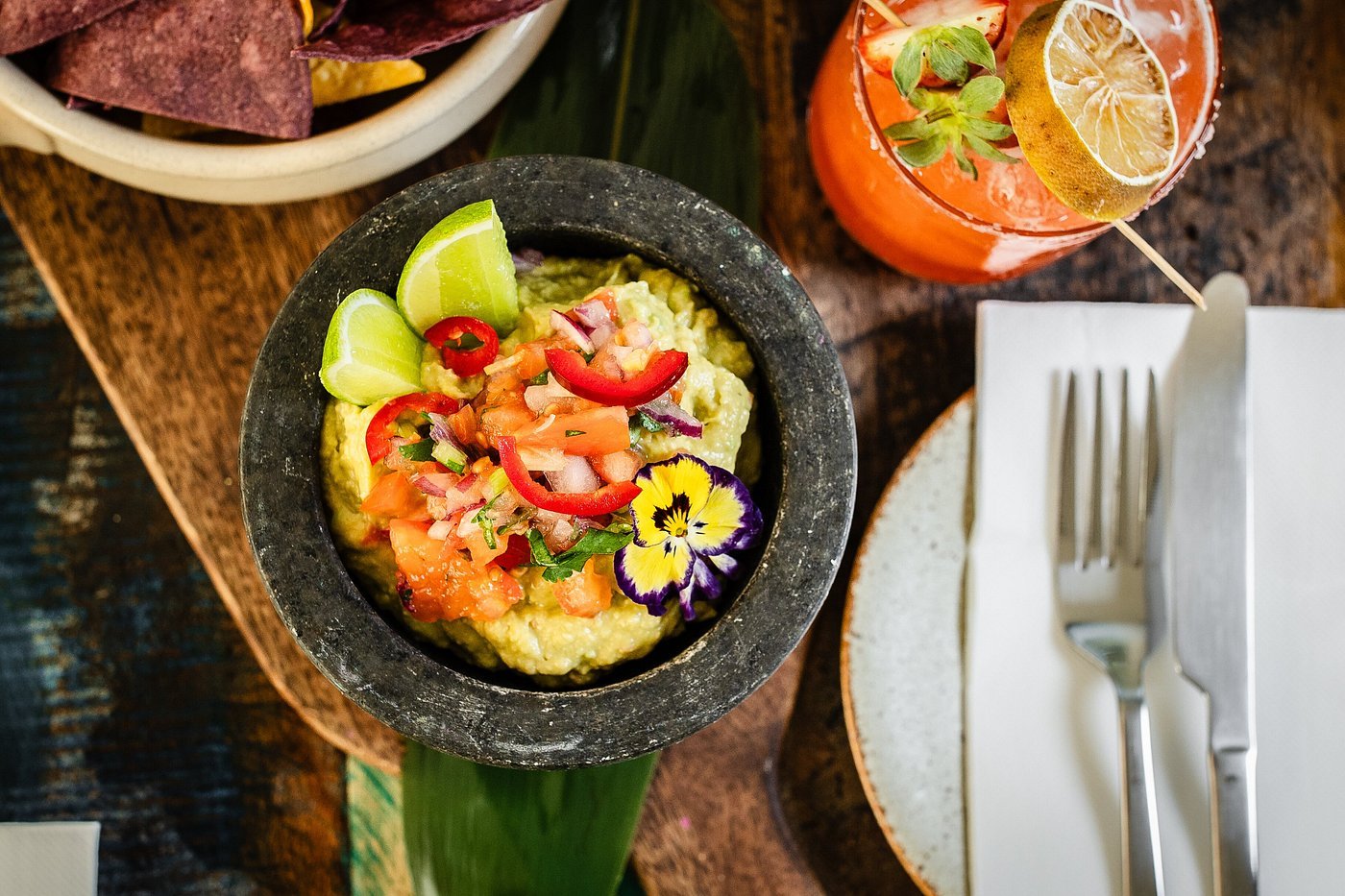 A bowl of guacamole topped with chopped tomatoes, red onions, cilantro, sliced red chili peppers, and lime wedges. A purple and yellow edible flower is on the side of the guacamole. To the right, there's a cocktail garnished with lemon slices and her
