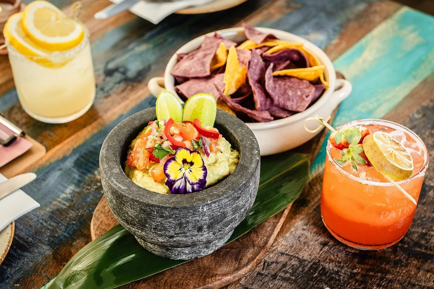 Table with Mexican food including guacamole with lime wedges, tortilla chips, and pink cocktail with lemon slices.