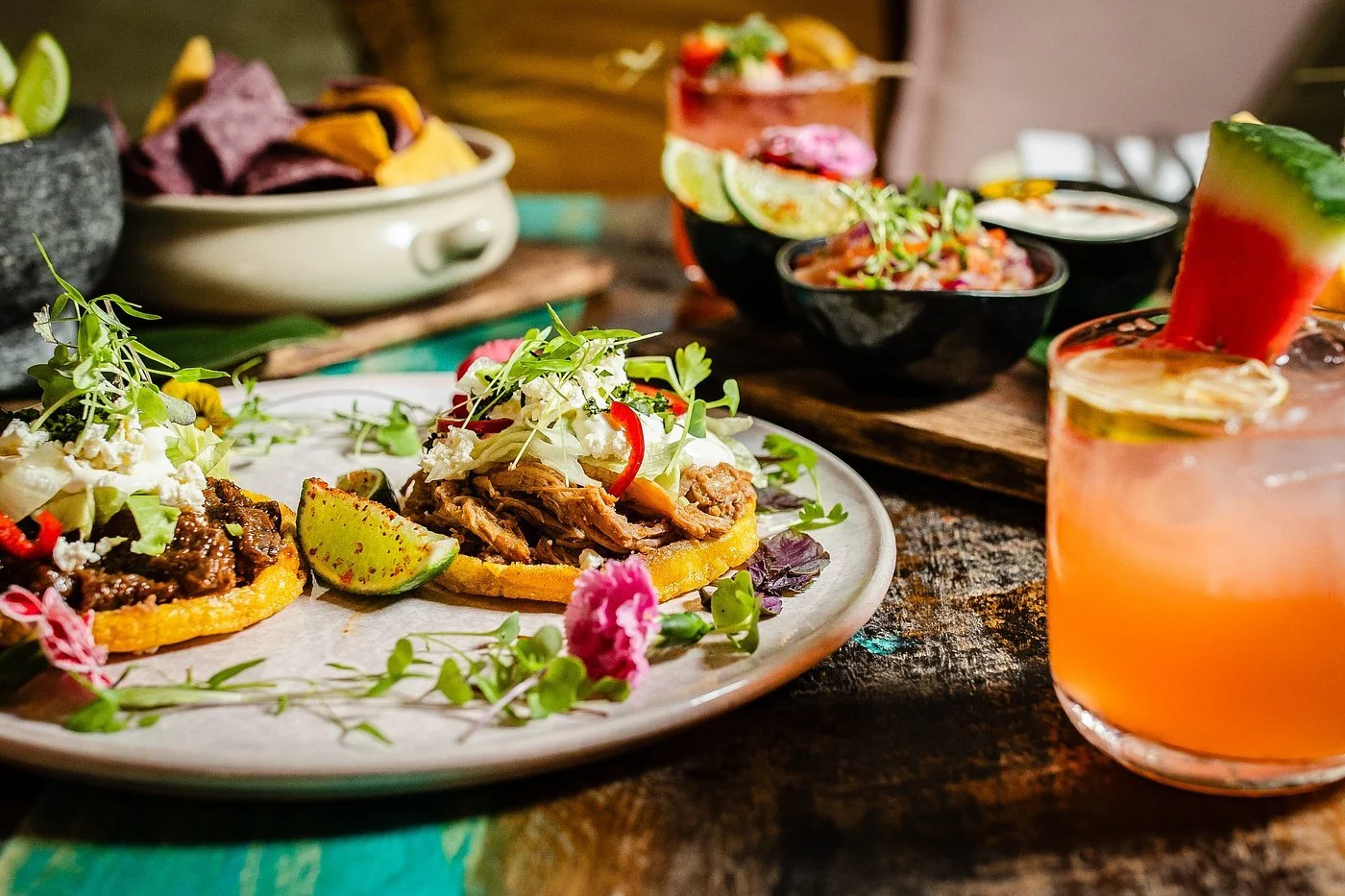Mexican dishes including tacos with shredded meat and garnishes, assorted salsas in small bowls, watermelon drink with lime, on a rustic wooden table.