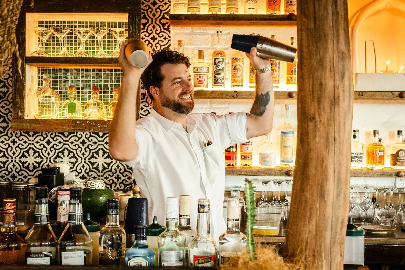 A smiling man with a beard and tattoos is shaking two cocktail shakers inside a bar. There are various bottles of alcohol and bar tools on the counter with a decorative backsplash and shelves filled with liquor bottles in the background.
