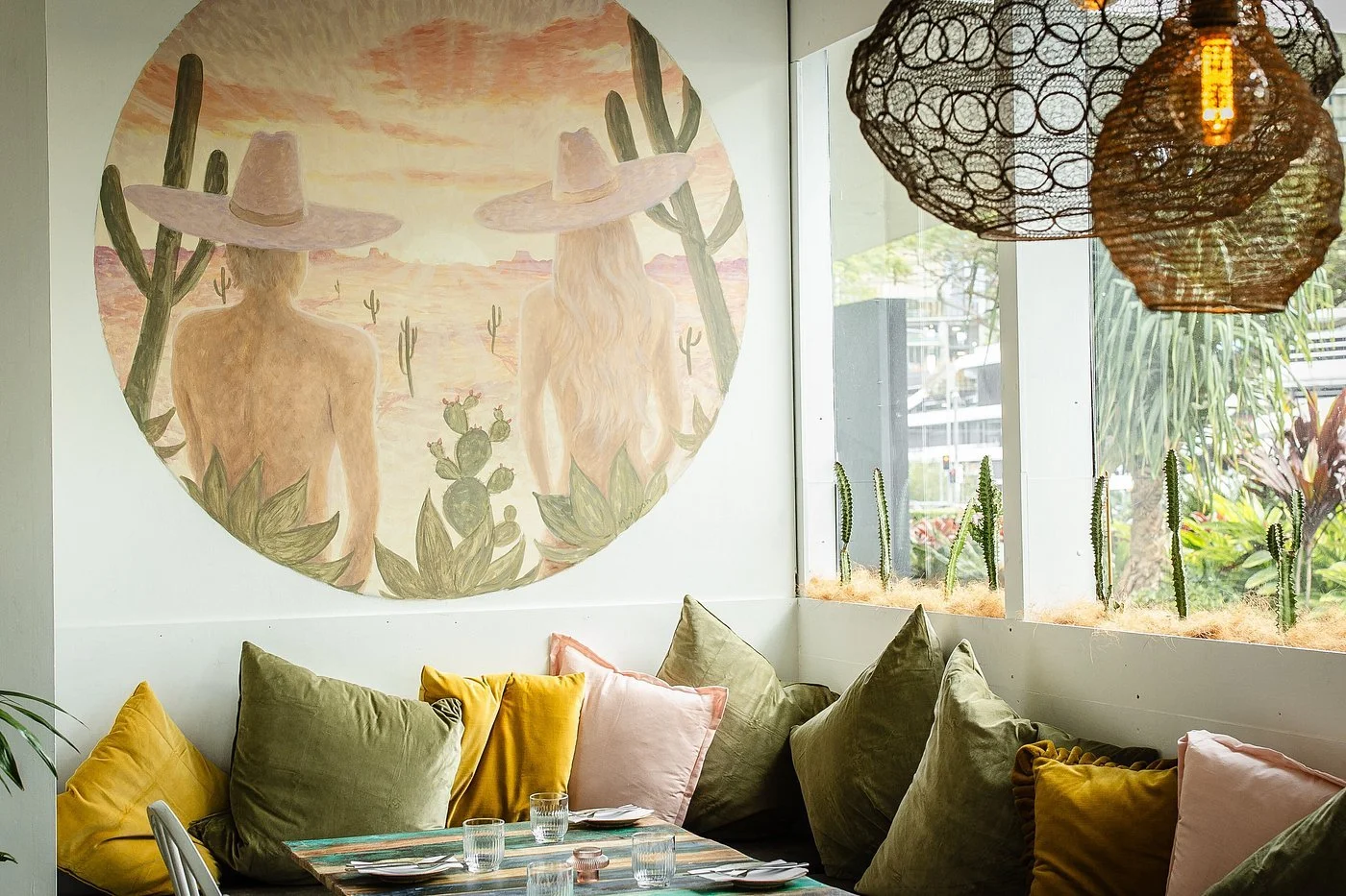 A cozy restaurant corner with a pastel-colored mural of a desert scene featuring two nude figures wearing large hats, surrounded by cacti. The same scene repeats on a circular canvas. The space is decorated with hanging woven lanterns, large cushions