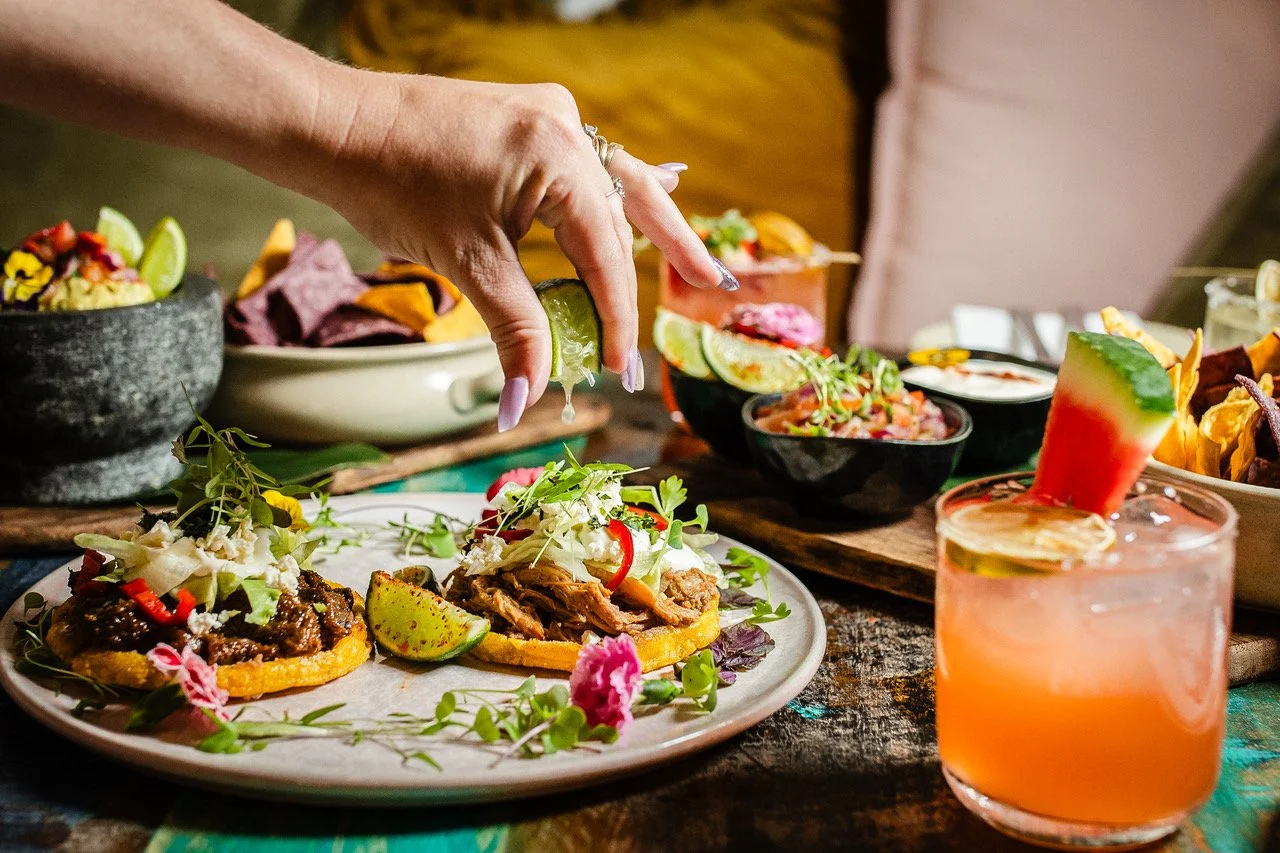 Person garnishing tacos with lime and microgreens on a rustic wooden table with bowls of salsa, chips, and cocktails.