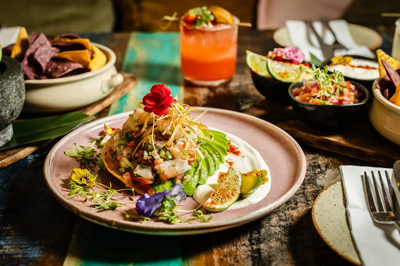 A plate of chopped salad garnished with flowers and lime wedges, surrounded by various bowls of Mexican dishes and a cocktail on a rustic wooden table.
