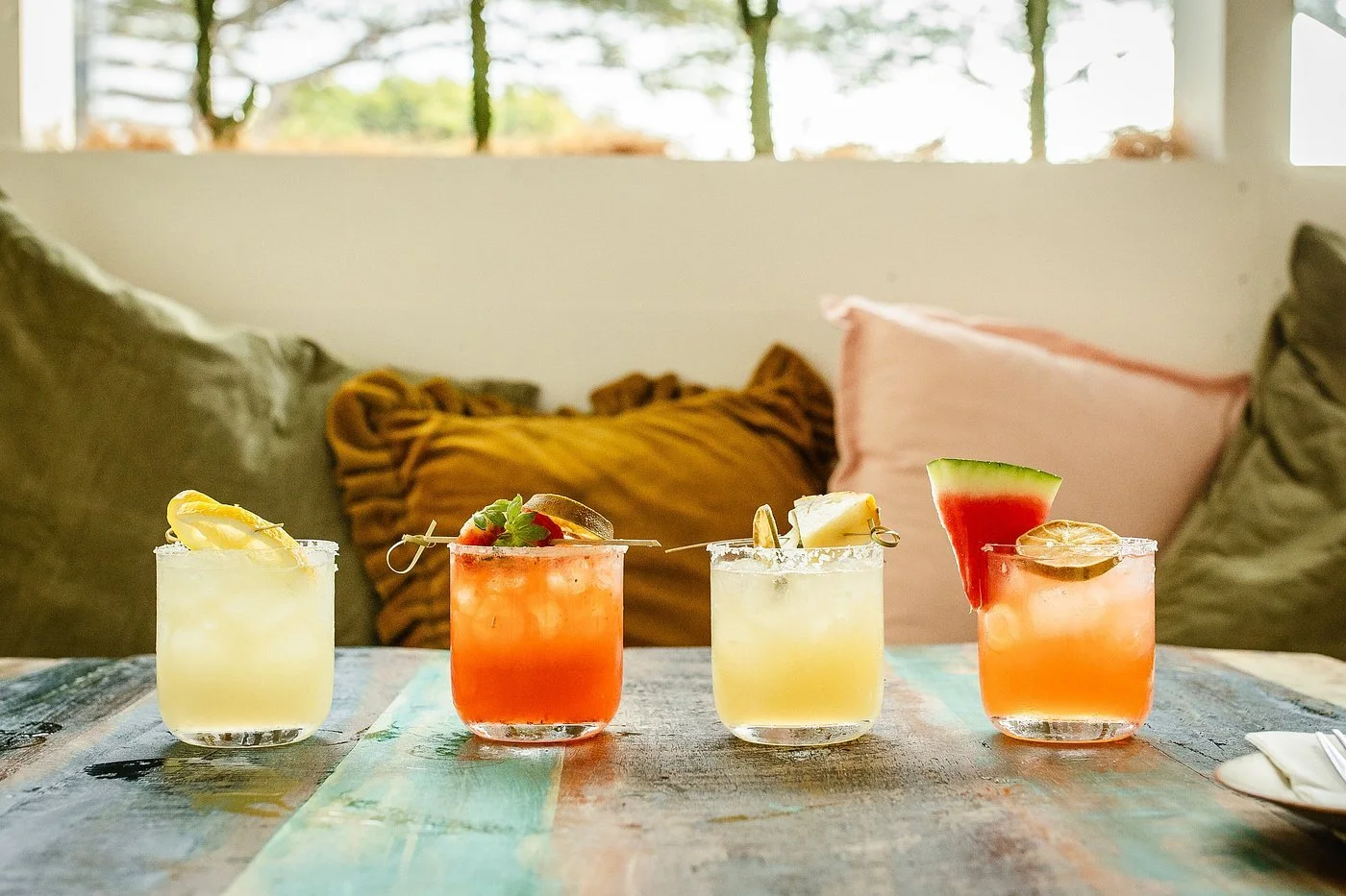 Four colorful cocktails with garnishes on a table in front of cushions and pillows on a couch.