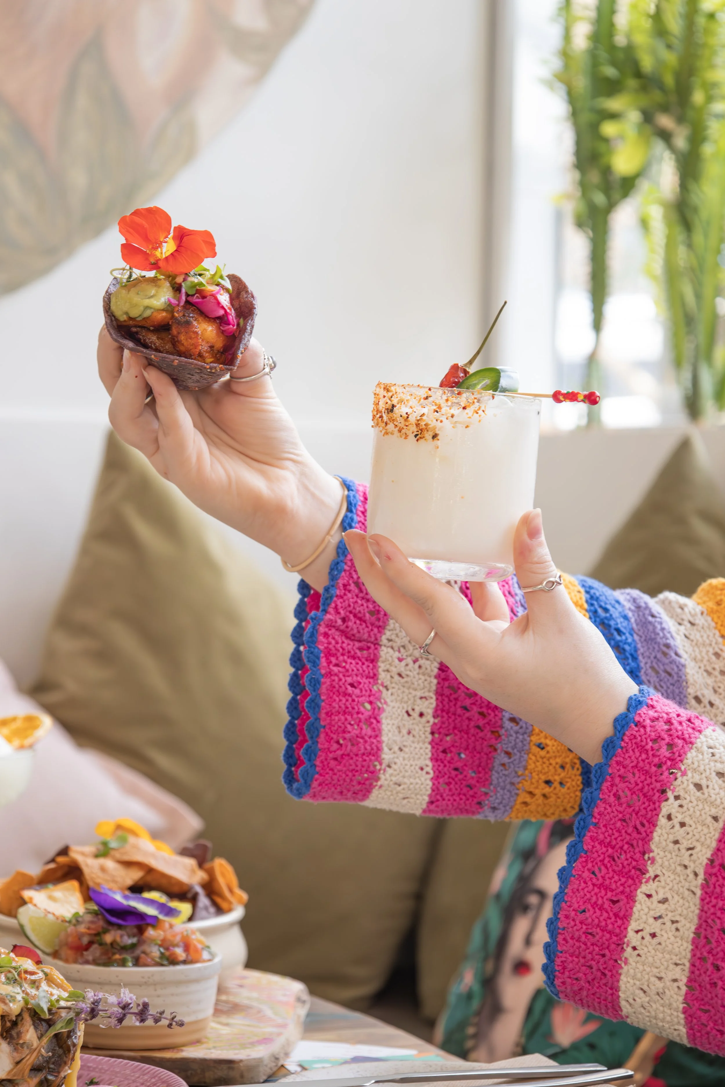 Person holding a cocktail garnished with fruit and spicy seasoning in one hand and a taco filled with toppings in the other, with colorful plates of food on the table.