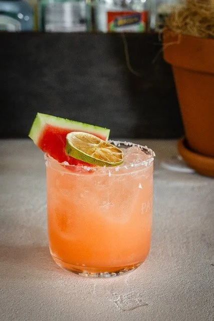 Pink cocktail in a glass garnished with a watermelon wedge and a slice of lime.