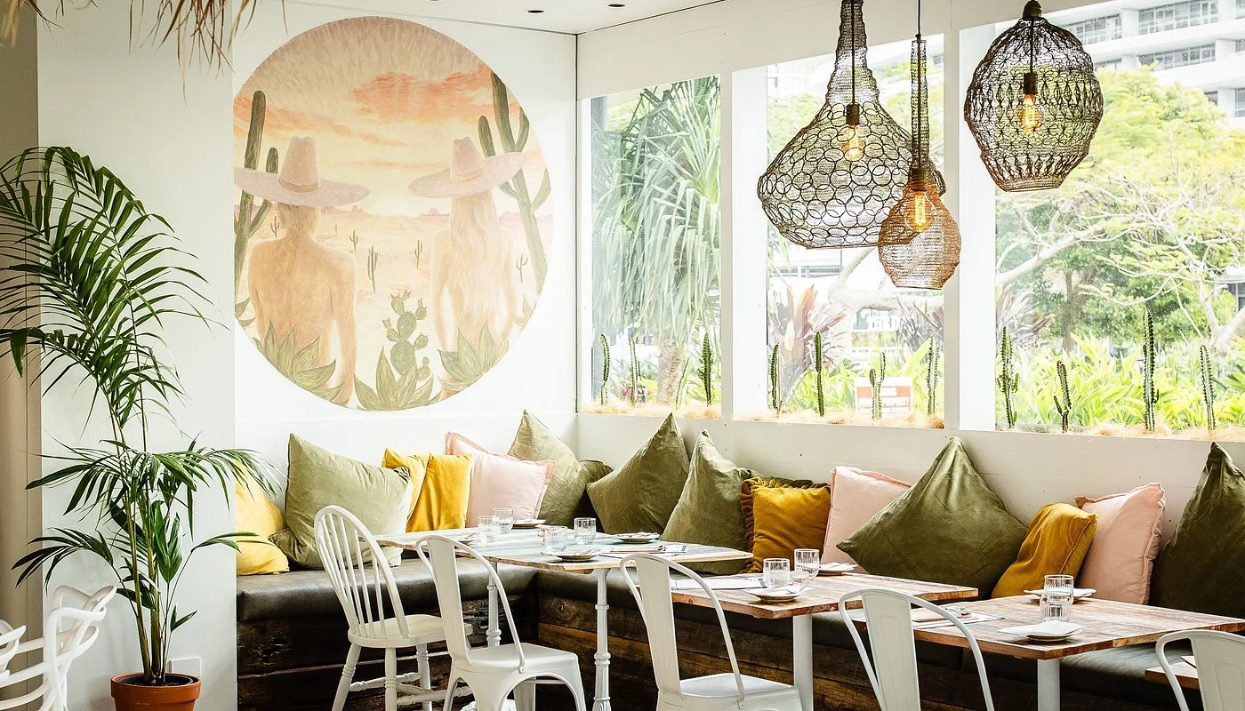 A cozy restaurant corner with a long cushioned bench lined with assorted pillows, tables set with glasses and plates, potted plants, and hanging woven pendant lights. A large window offers a view of trees and buildings outside, with a wall art featuring two women wearing sombreros in a desert scene.