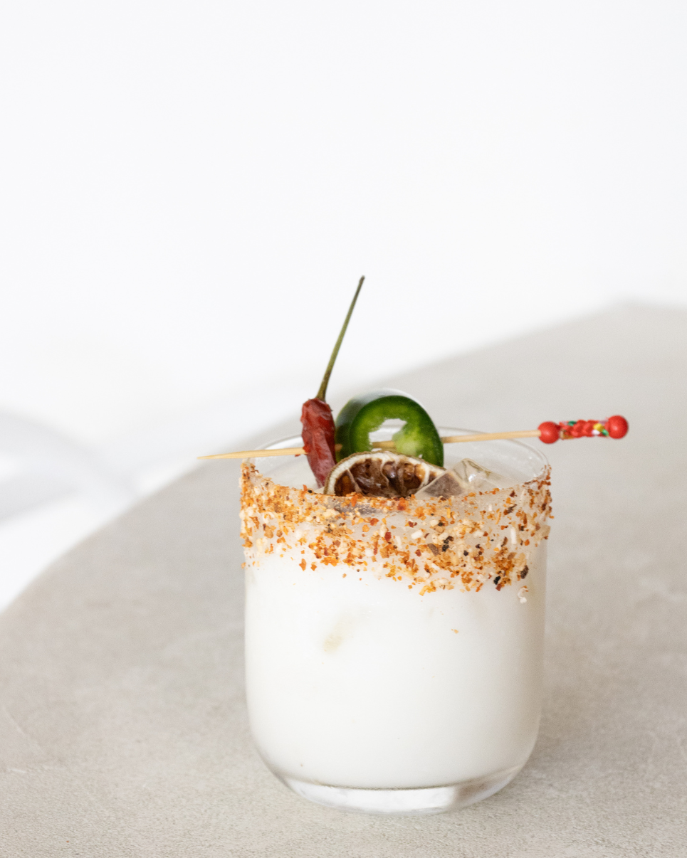 A cocktail garnished with jalapeño, chili pepper, and a spicy skewer, rimmed with chili powder or seasoning on a white surface.