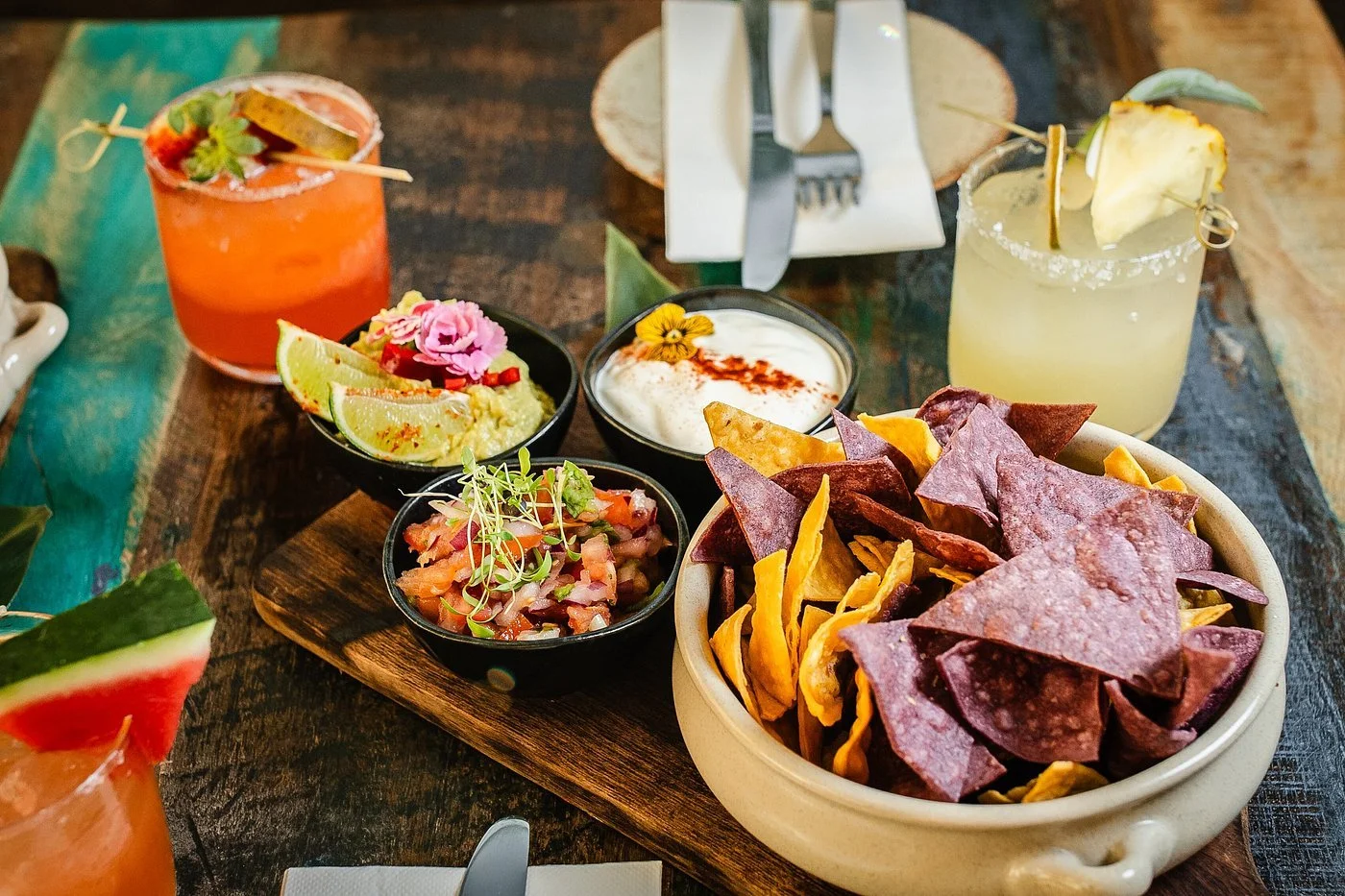 A wooden table with bowls of colorful snacks, including tortilla chips, salsa, guacamole, and sour cream, along with two cocktails garnished with fruit slices and flowers.