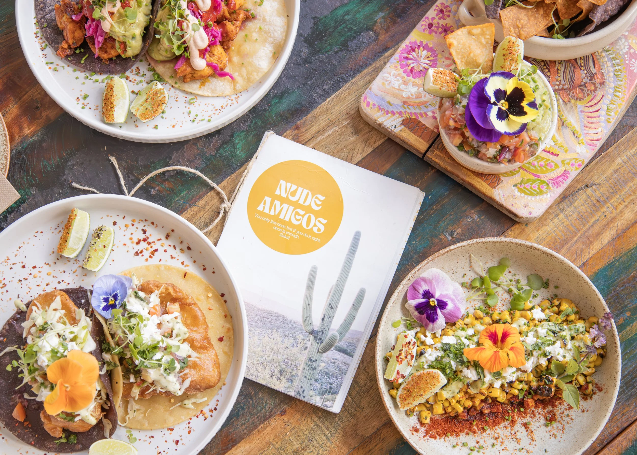 A variety of Mexican dishes on black plates, including tacos with meat, vegetables, and garnishes, burritos, a bowl of chips with guacamole, small bowls of salsa, and other Mexican food.