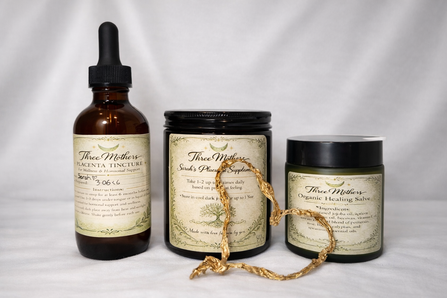 Three Mothers branded wellness products including a placenta tincture in a dropper bottle, a placenta supplement in a jar with a gold necklace draped over it, and an organic healing salve in a jar with a black lid, all on a white surface.