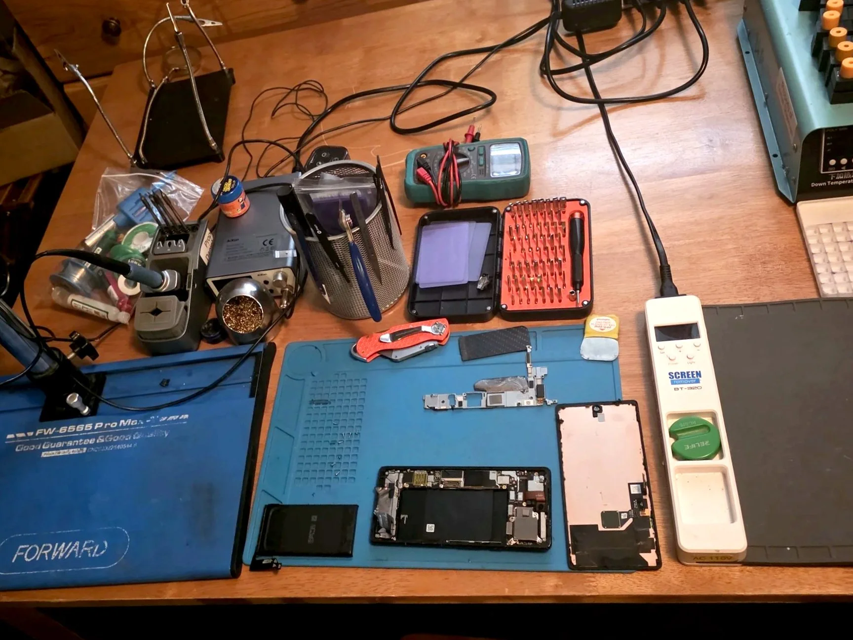 Disassembled smartphone with tools, screwdriver set, multimeter, and soldering equipment on a wooden workbench.