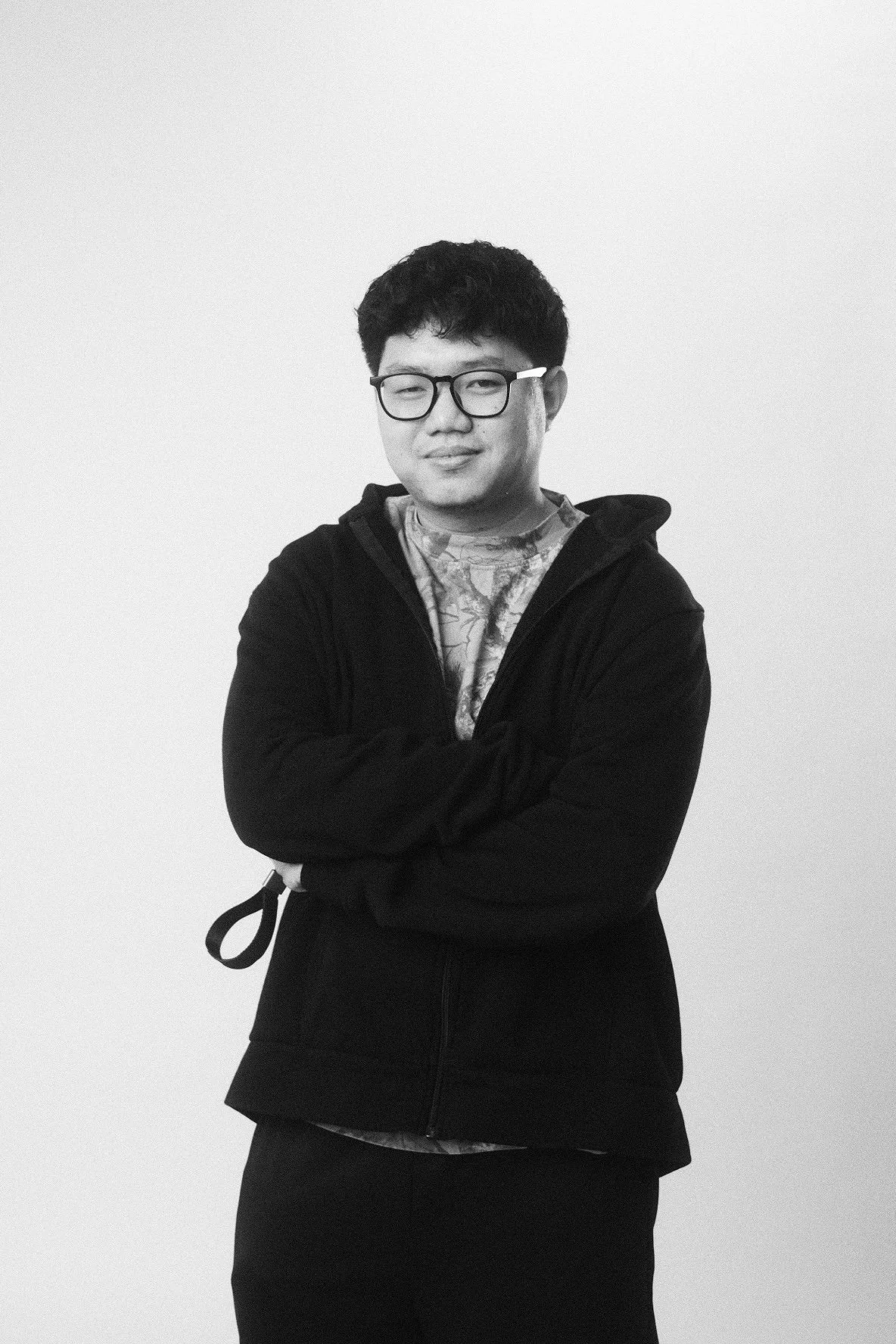 A young man with glasses, wearing a patterned shirt and a black zip-up jacket, standing against a plain white background.