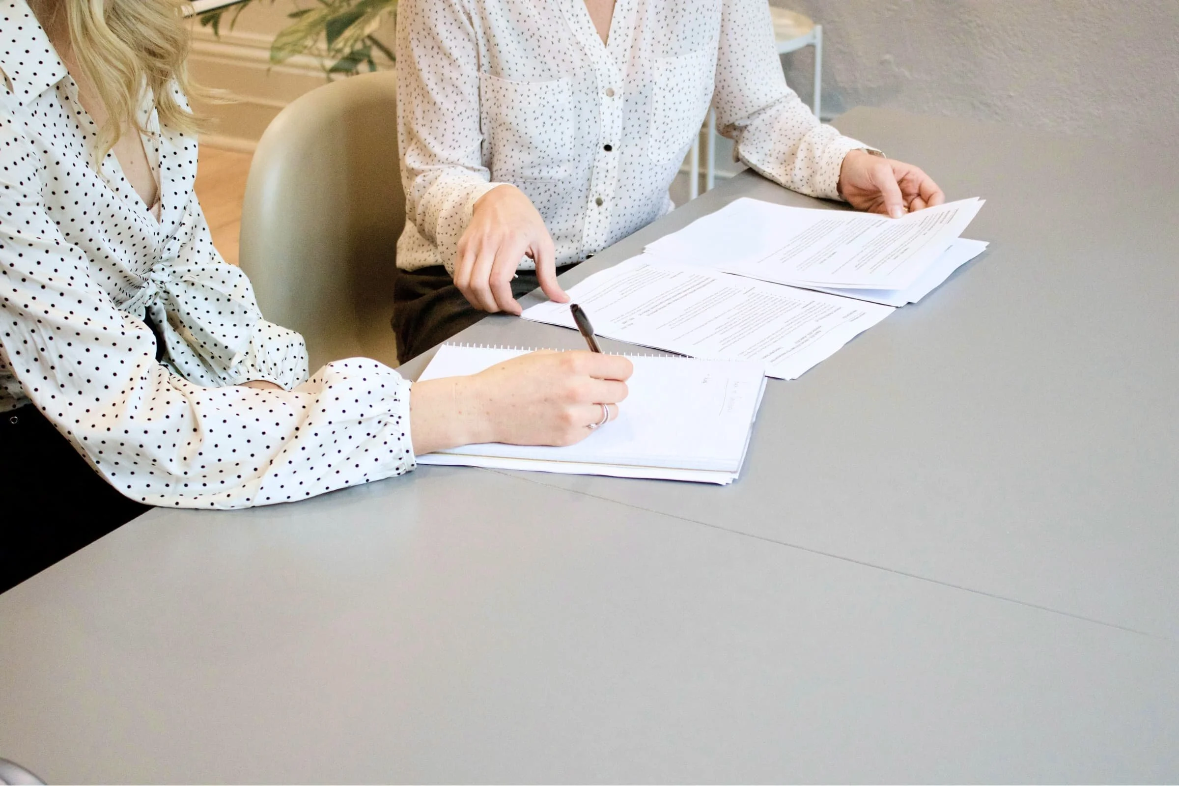 Two women sitting at a table with documents; one woman is pointing at the papers with her finger, the other is writing notes.