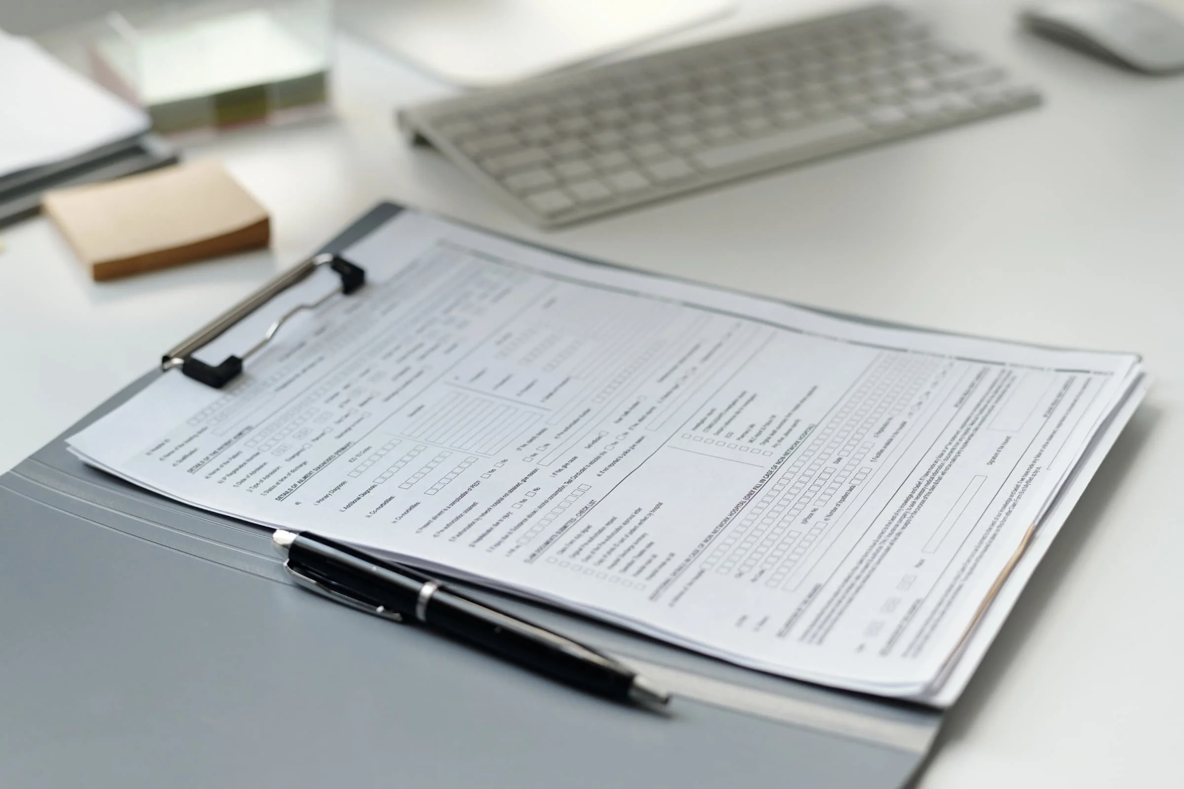 Office desk with a clipboard holding printed forms, keyboard, notepad, and other office supplies.
