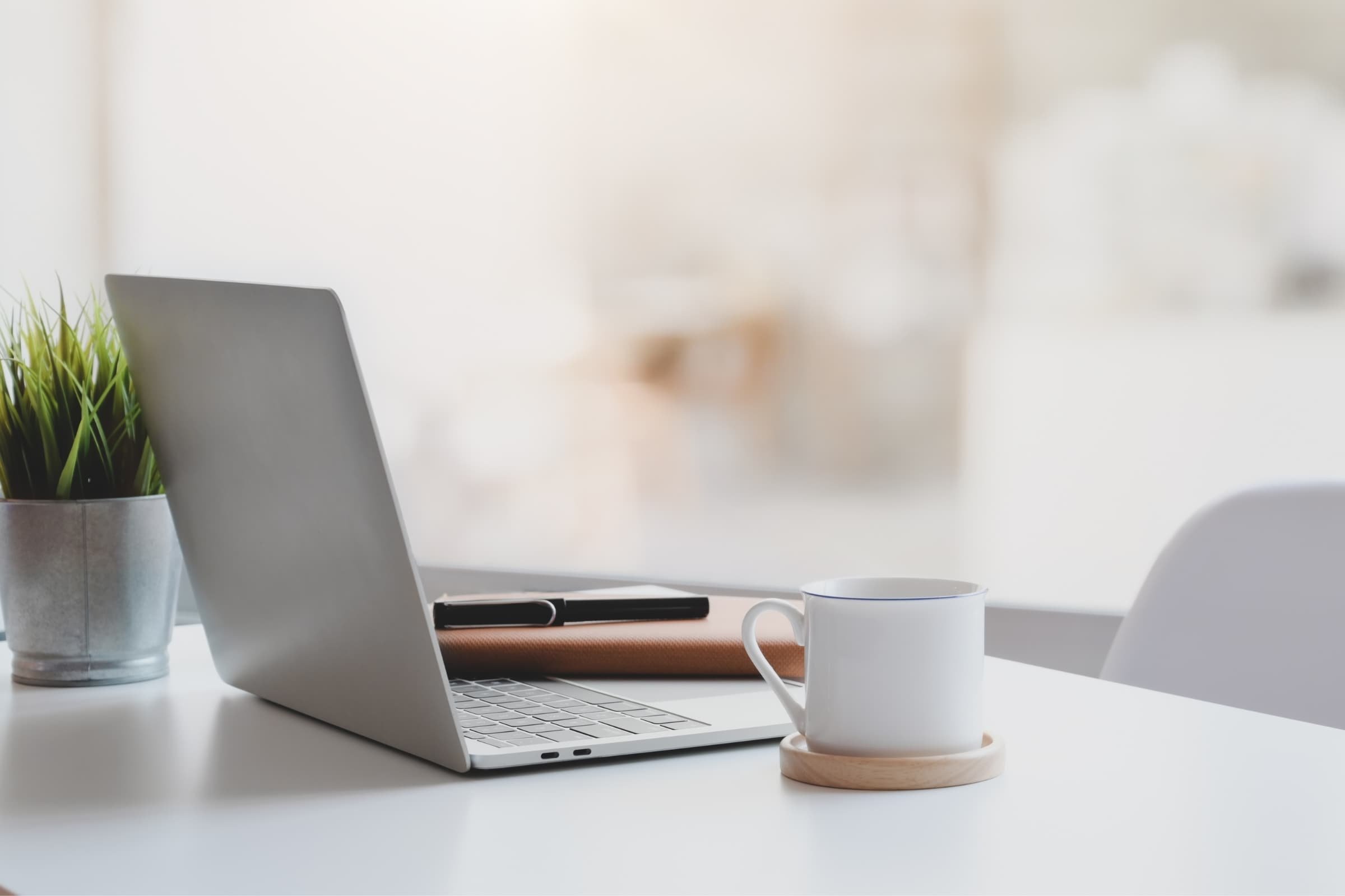 A laptop on a white desk with a potted plant on the left, a black notebook behind the laptop, a white coffee mug on a wooden coaster on the right, and a pen on a cork pad, with a blurred background.