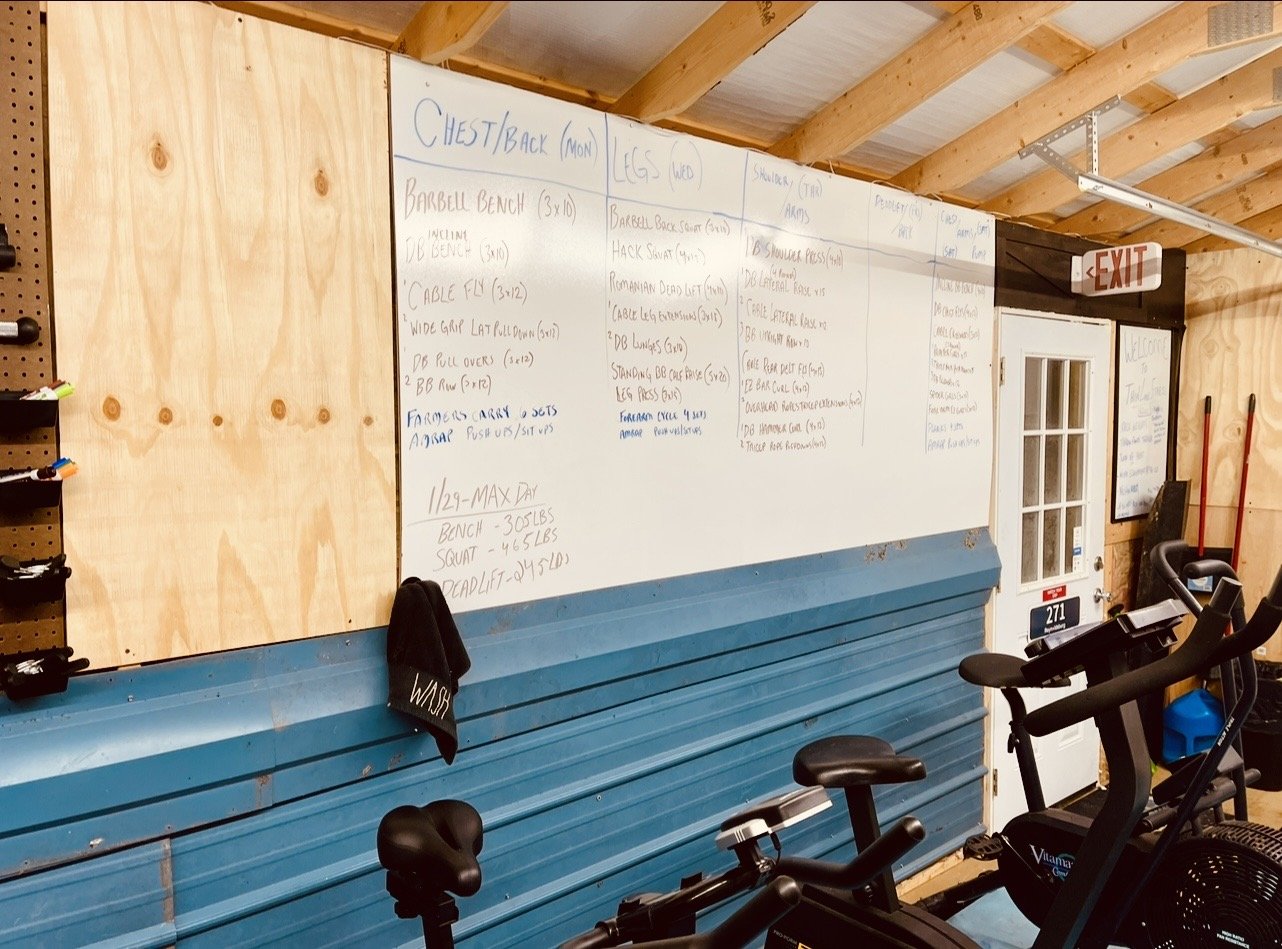 A gym interior with a whiteboard on a wooden wall containing workout routines and notes. There are exercise bikes in the foreground, a broom, and a door with an exit sign overhead.
