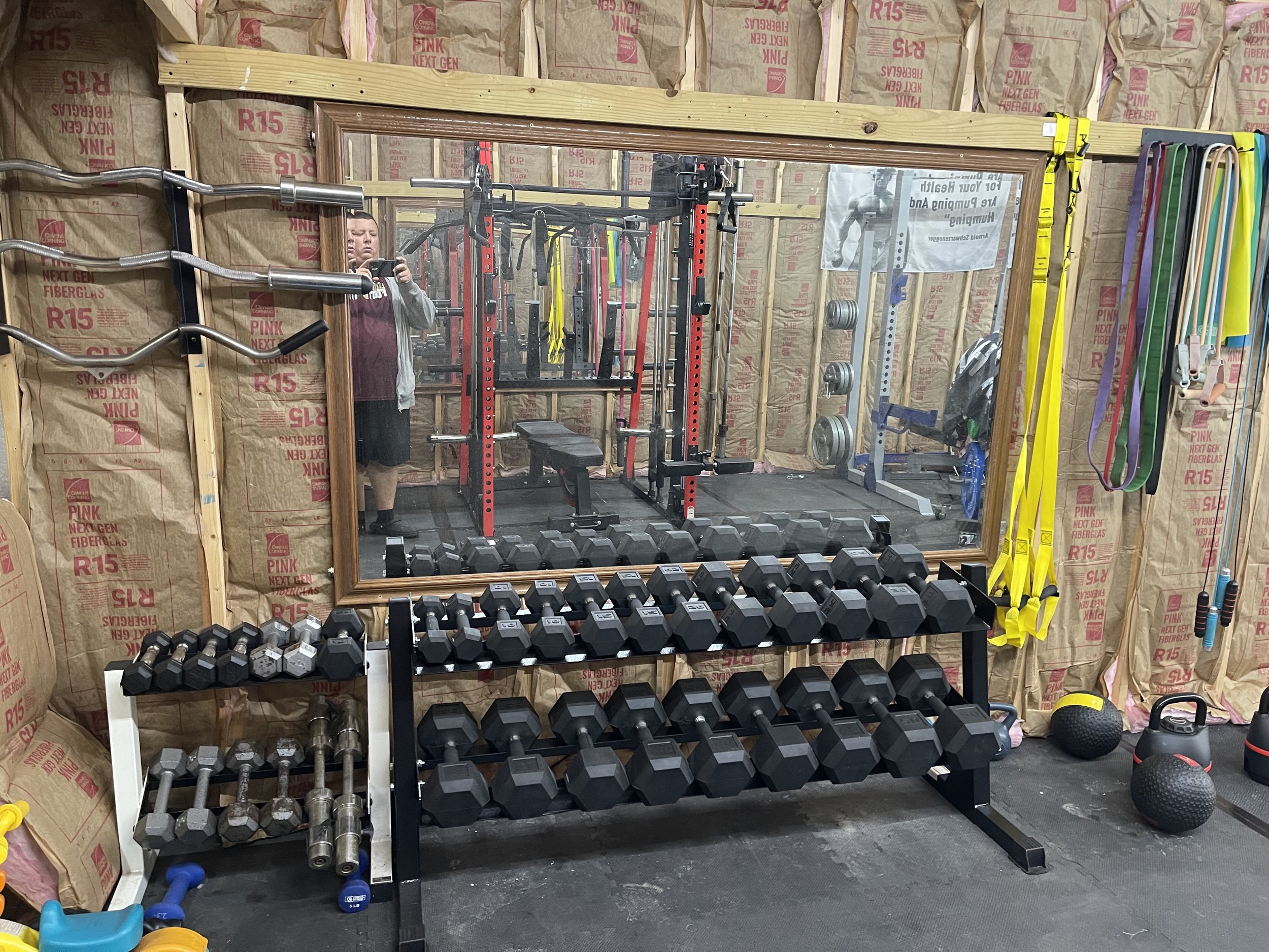 A home gym with a large mirror, dumbbell rack, a few kettlebells, foam rollers, and resistance bands attached to the wall, with an individual taking a photograph of the gym.