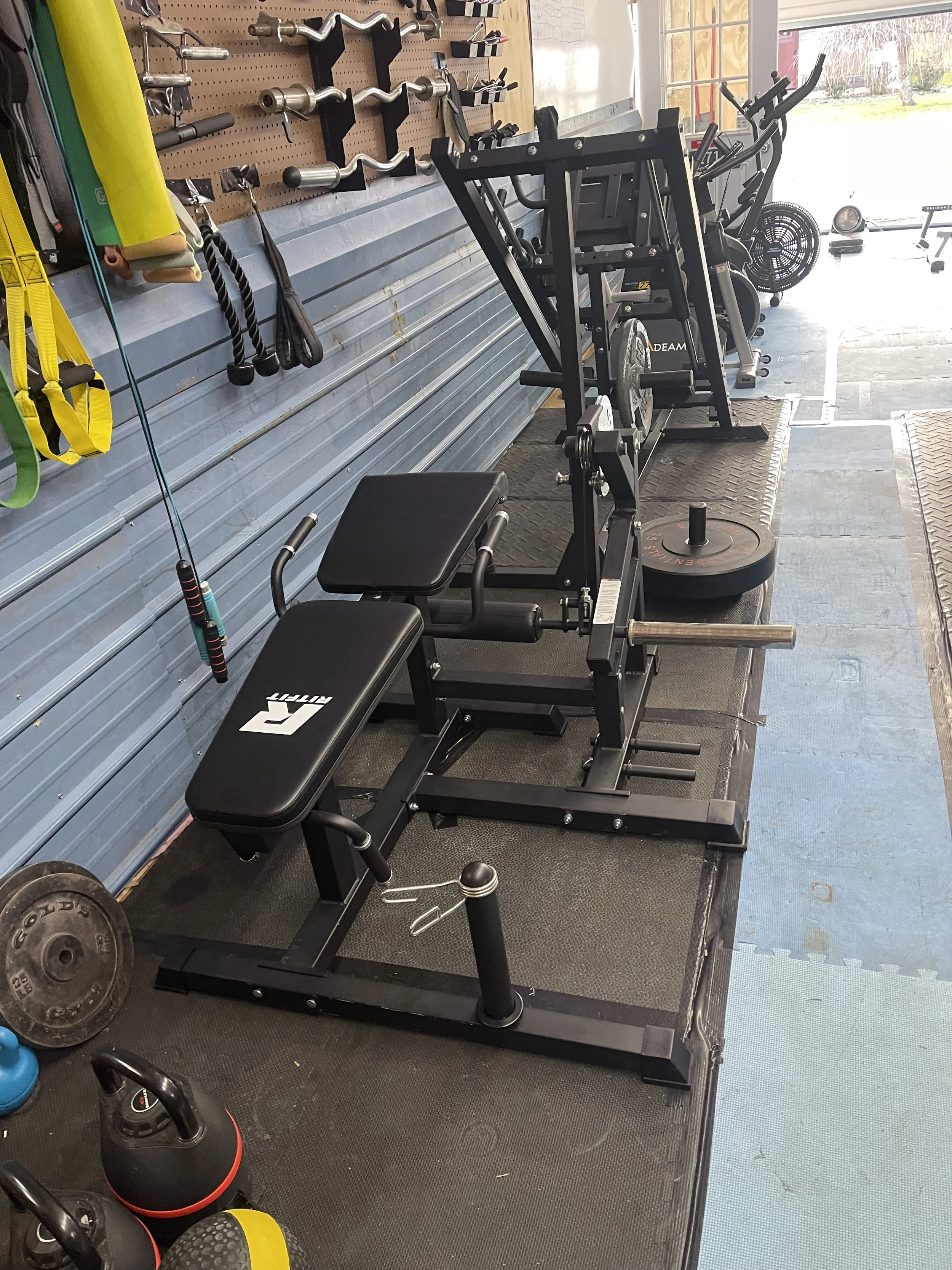A home gym with various exercise equipment, including a weight bench, a rowing machine, a black weight plate, kettlebells, and resistance bands. The wall has hooks holding tools and accessories, with a window showing outdoor scenery.