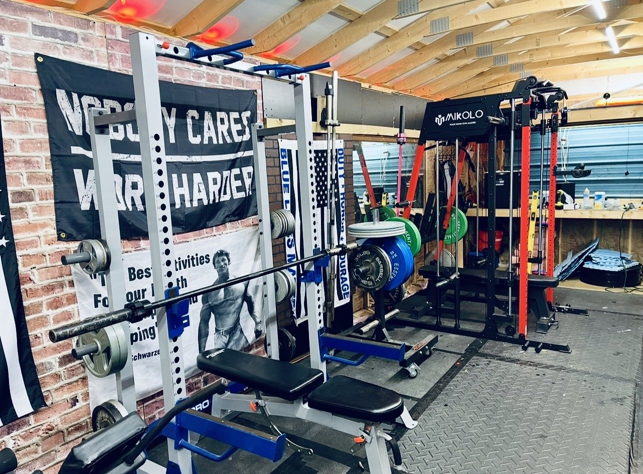 Home gym with weightlifting equipment, barbells, weight plates, resistance bands, and a pull-up station. Exposed brick wall and wooden ceiling.