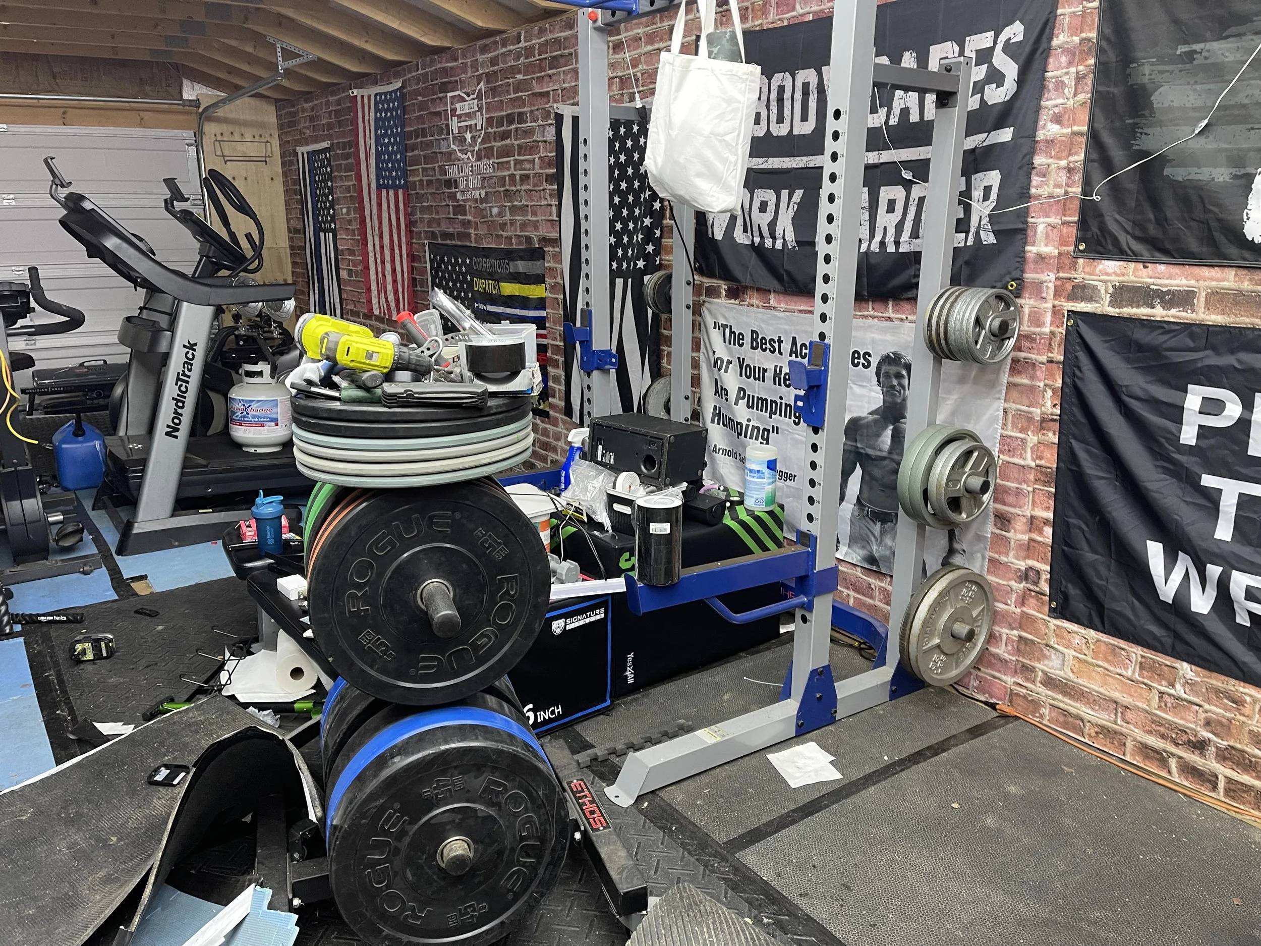 Home gym with a treadmill, weight plates, energy drinks, and fitness posters and flags on brick wall.