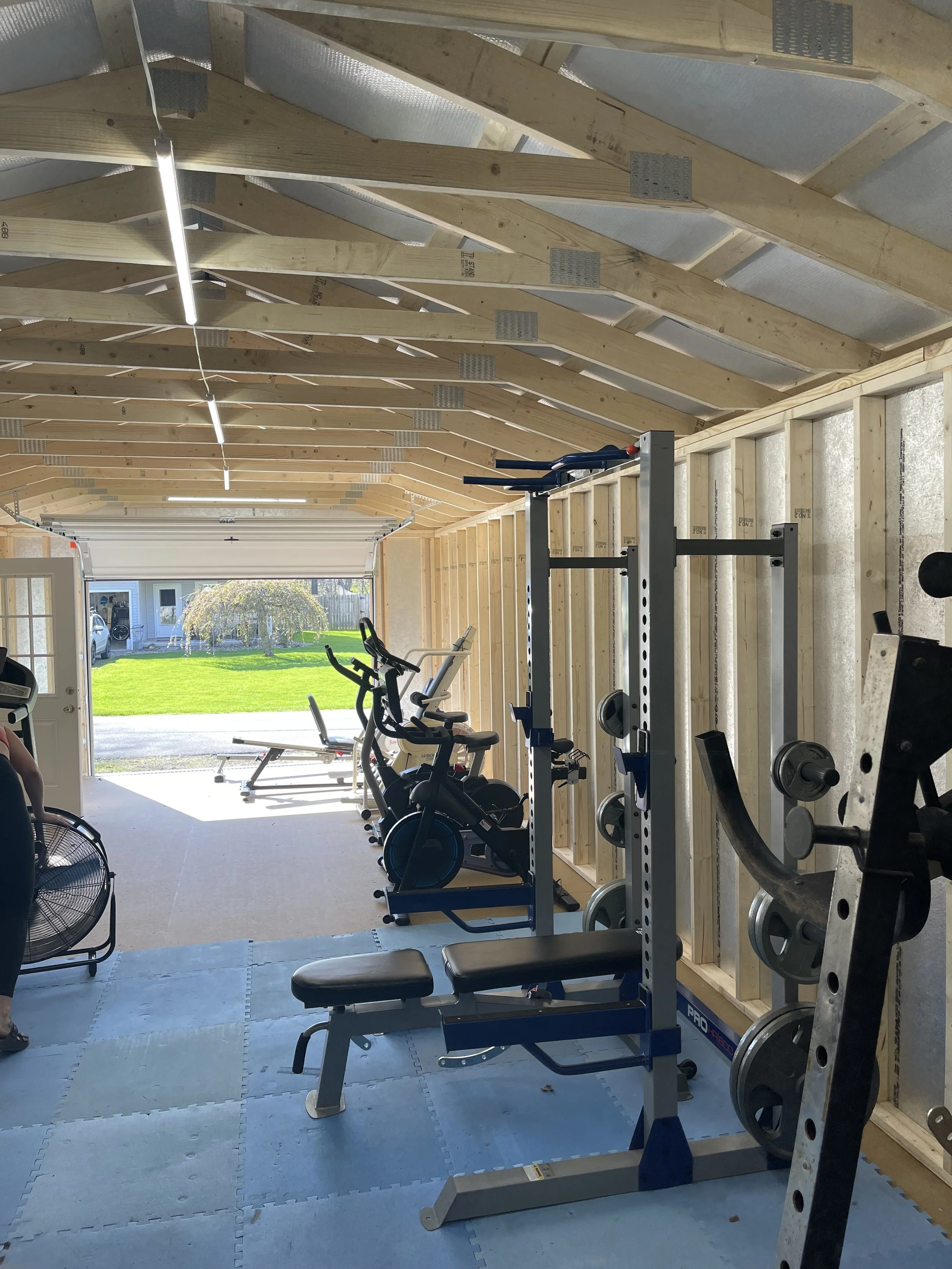 Home gym with weight rack, exercise bikes, bench, and outdoor view through open garage door, with wood framing and unfinished ceiling.
