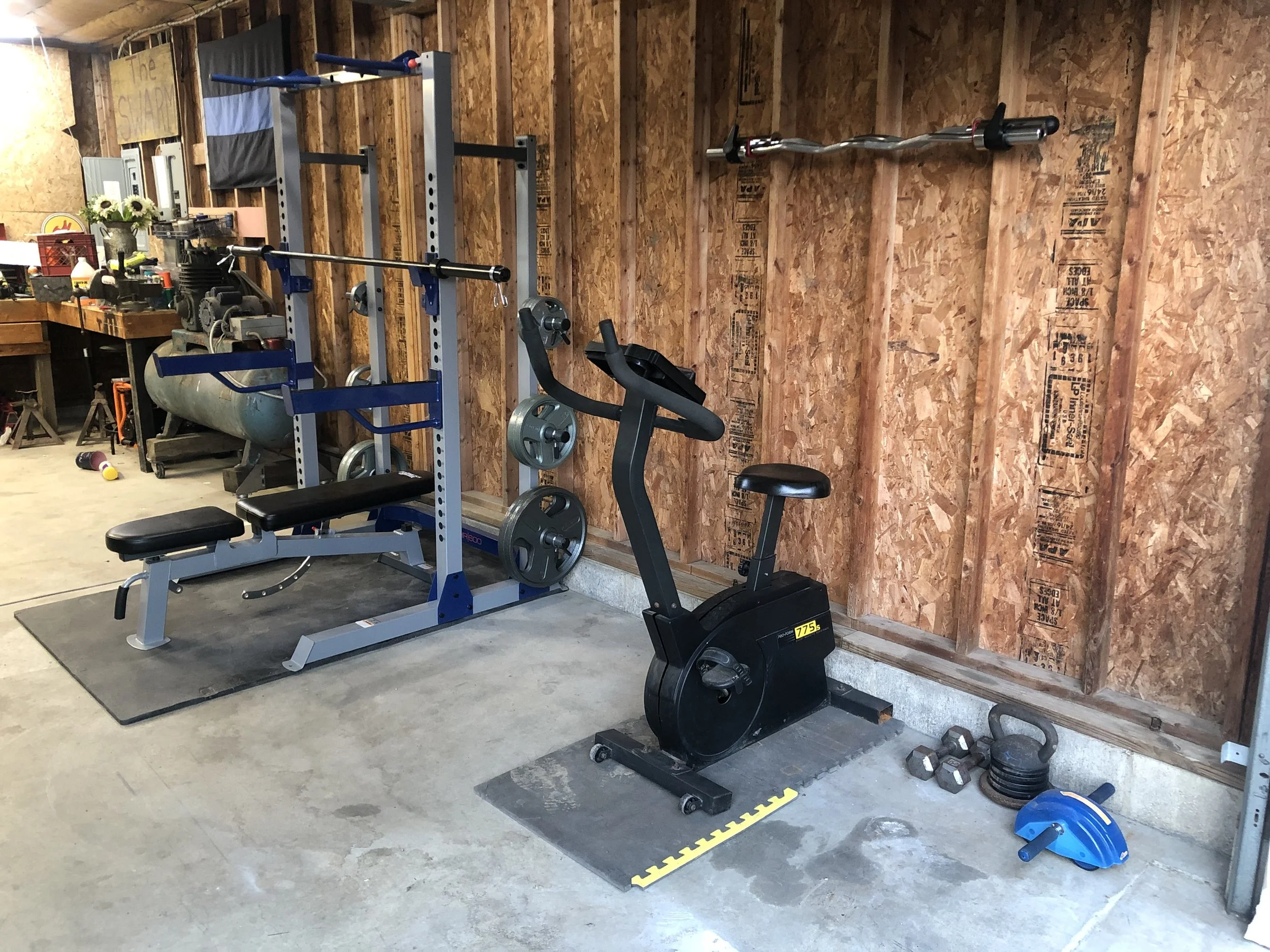 Home gym setup with weightlifting bench, barbell, weight plates, stationary bike, dumbbells, kettlebell, and a wall-mounted barbell in a garage with wooden walls.