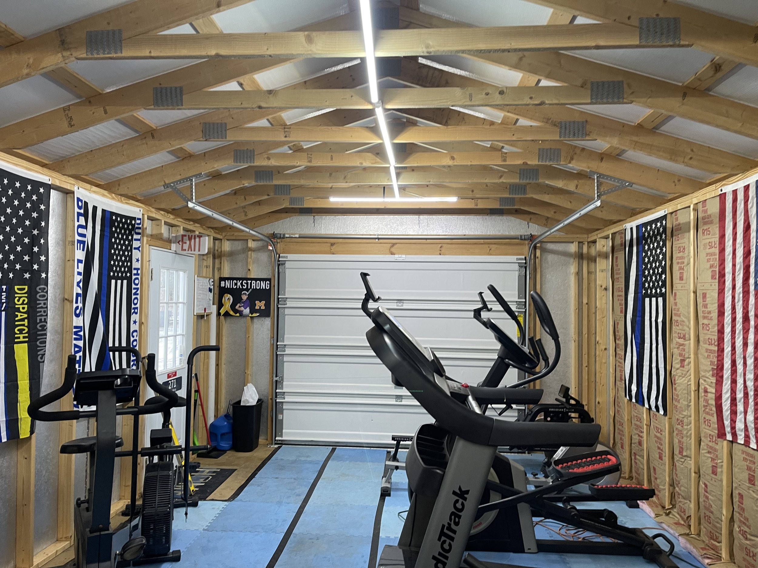 A home gym with an exercise bike and elliptical machine, with Minnesota Vikings flags, a Nick Strong banner, a mop, and cleaning supplies, in a garage with unfinished walls and a garage door.