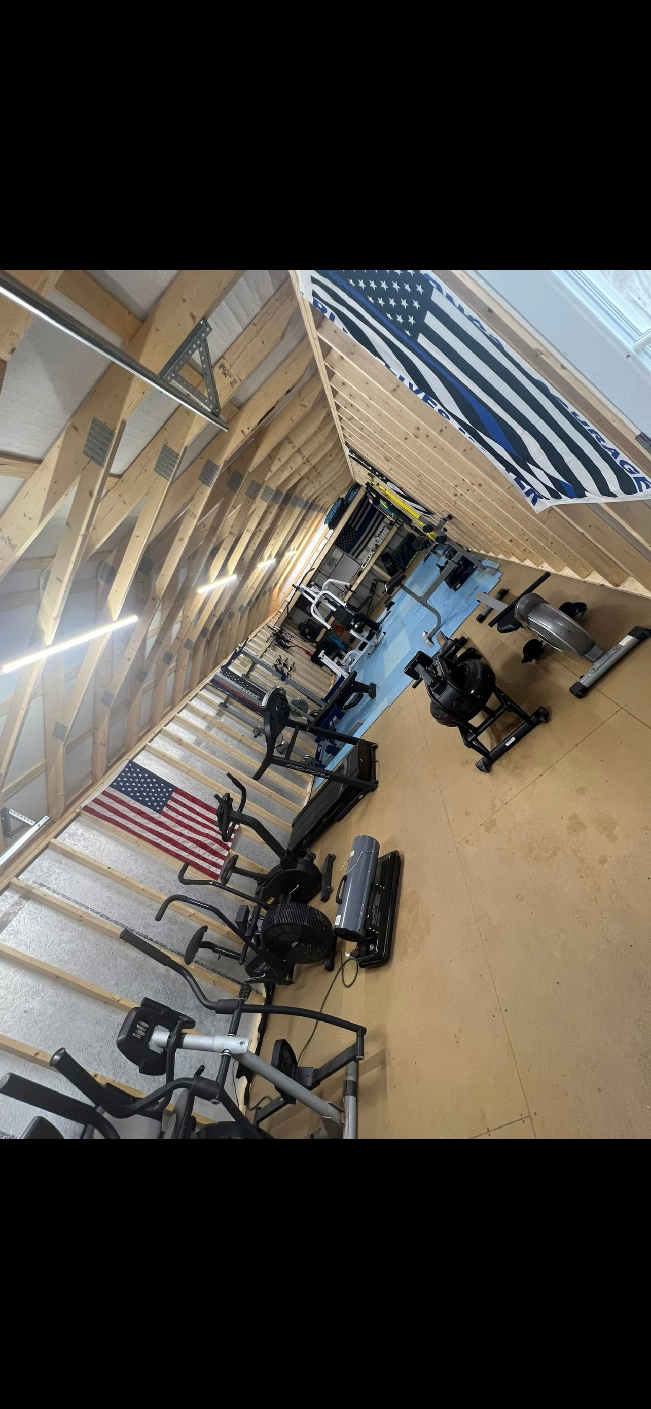 A home gym with various workout equipment such as treadmills, ellipticals, and weights, set against unfinished wooden walls with an American flag banner hanging from the ceiling.