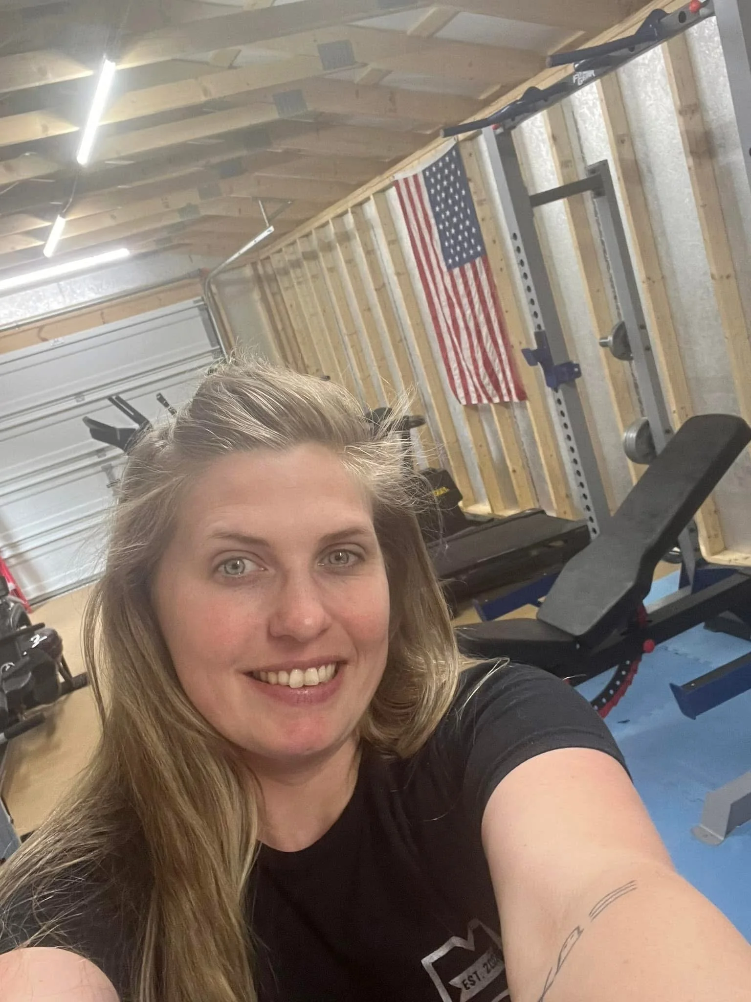 A woman taking a selfie in a home gym with wooden walls and an American flag hanging behind her. Gym equipment is visible, including a weight bench and pull-up bars.