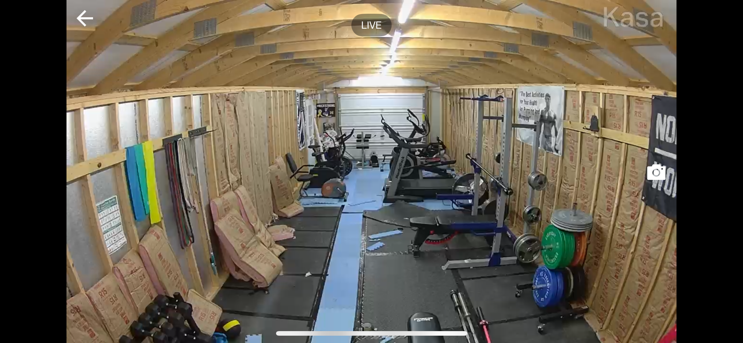 This image shows a home gym in a partially finished room with wooden framing and Insulation. The gym equipment includes exercise bikes, a treadmill, weight plates, and various tools and equipment for fitness training.