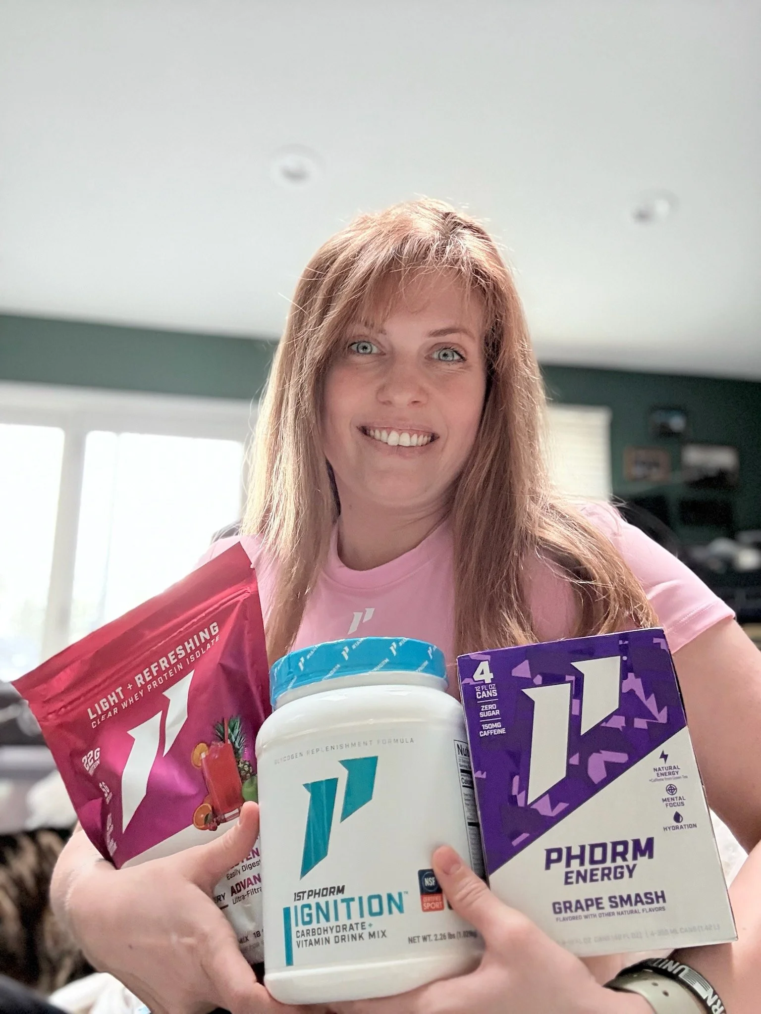 Woman holding various sports nutrition products in a room with large windows and a green wall.