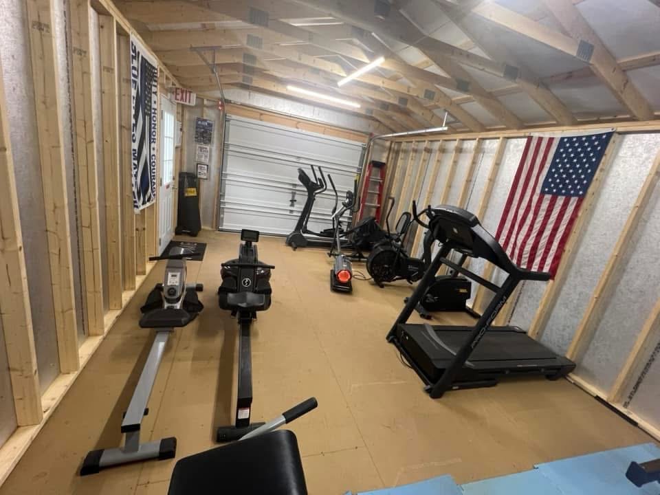 Home gym with treadmill, elliptical, rowing machine, and American flag on unfinished wall.