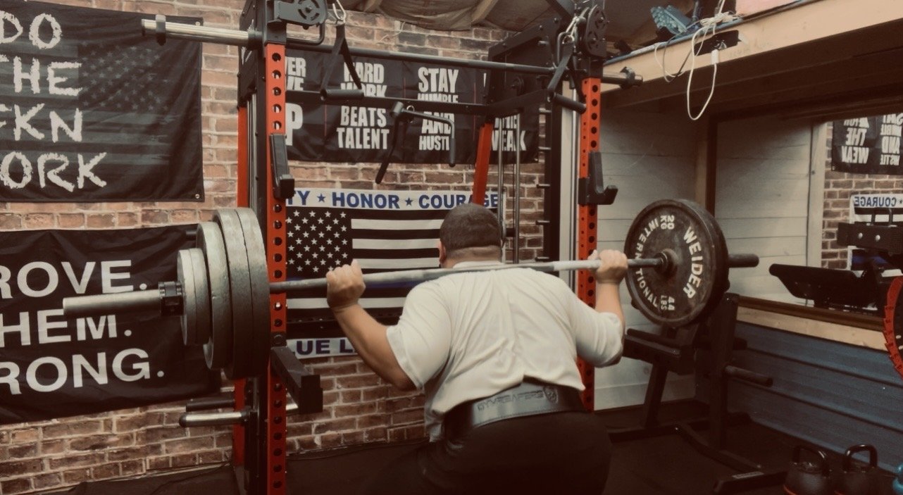 A man in a gym is performing a barbell squat with weights attached, standing with his back to the camera, facing a brick wall decorated with motivational posters and an American flag.