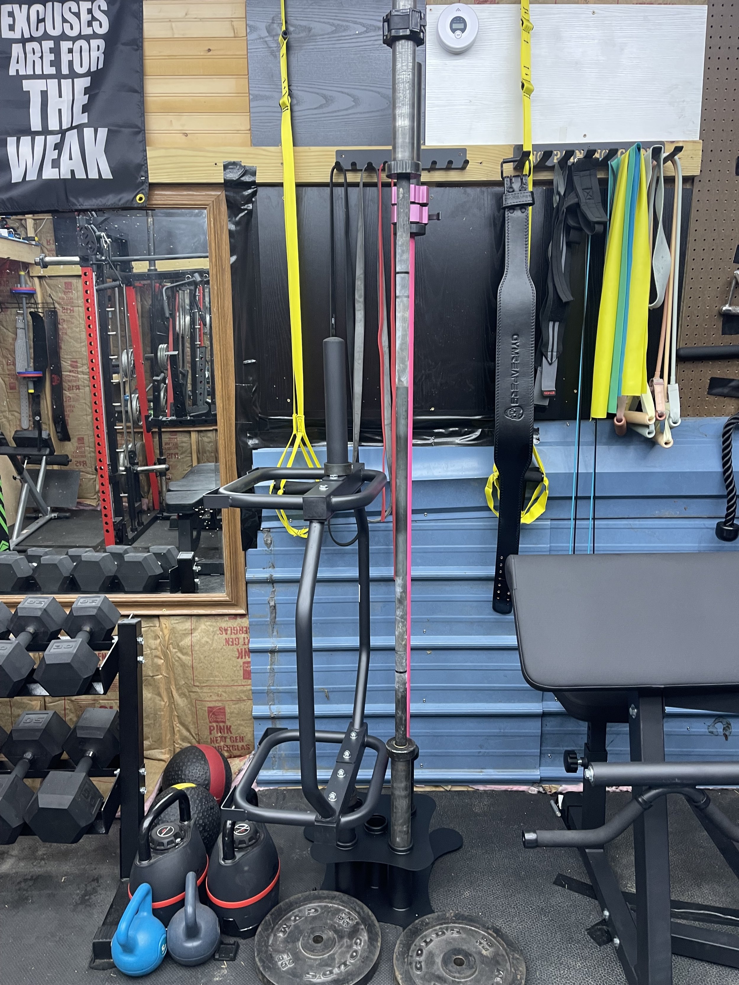 A gym wall with various fitness equipment including kettlebells, weights, resistance bands, and suspension trainers mounted on hooks, along with a mirror reflecting parts of the workout area.