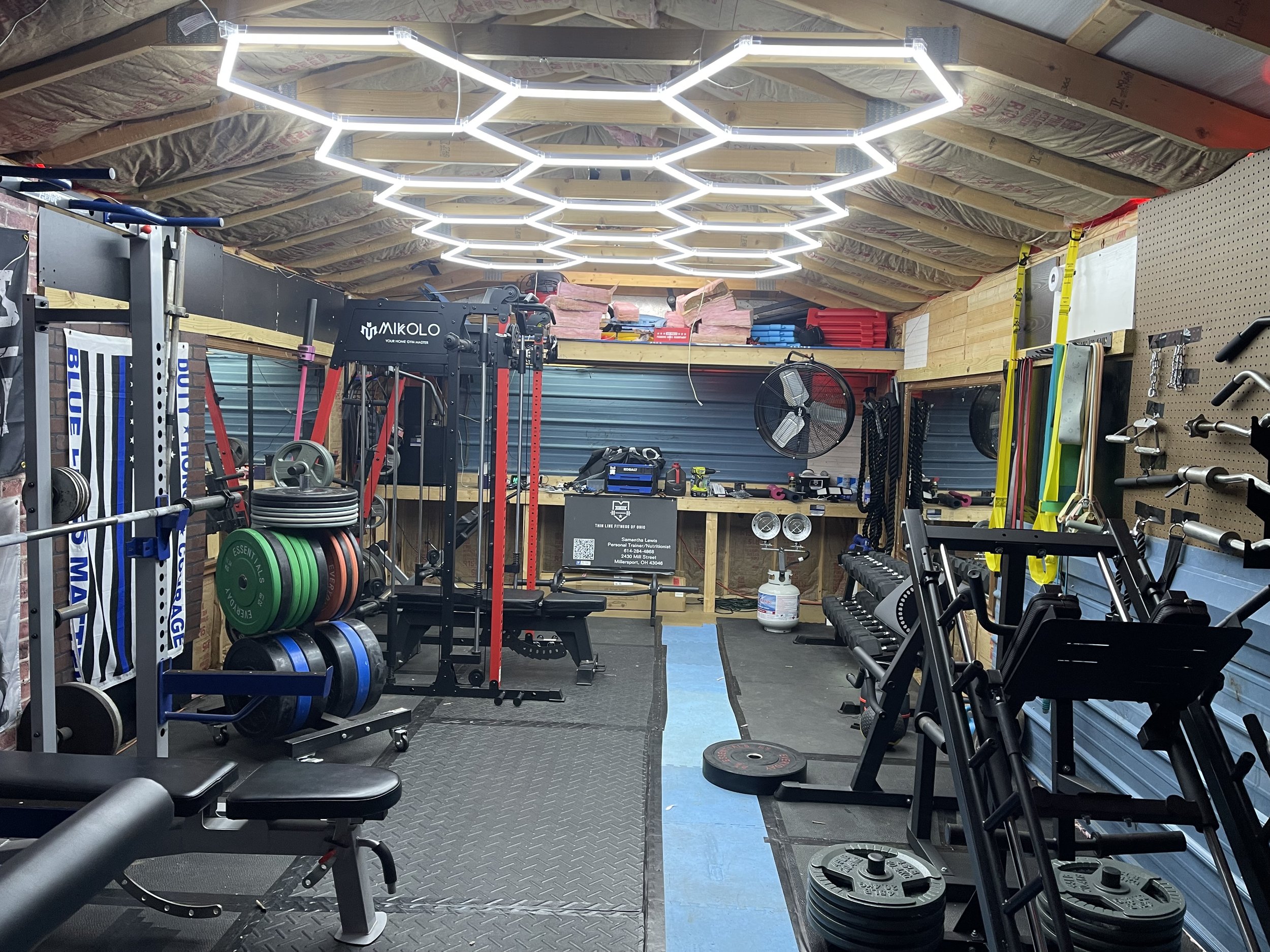 Home gym with weightlifting equipment, plates, dumbbells, and resistance bands, inside a garage with a hexagonal LED light fixture on the ceiling.