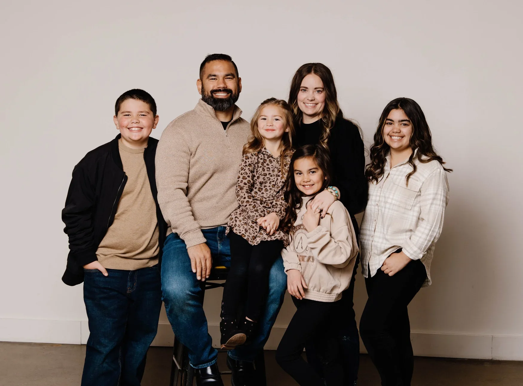 Family portrait of six people, including three children and two adults, posing together in front of a plain wall.