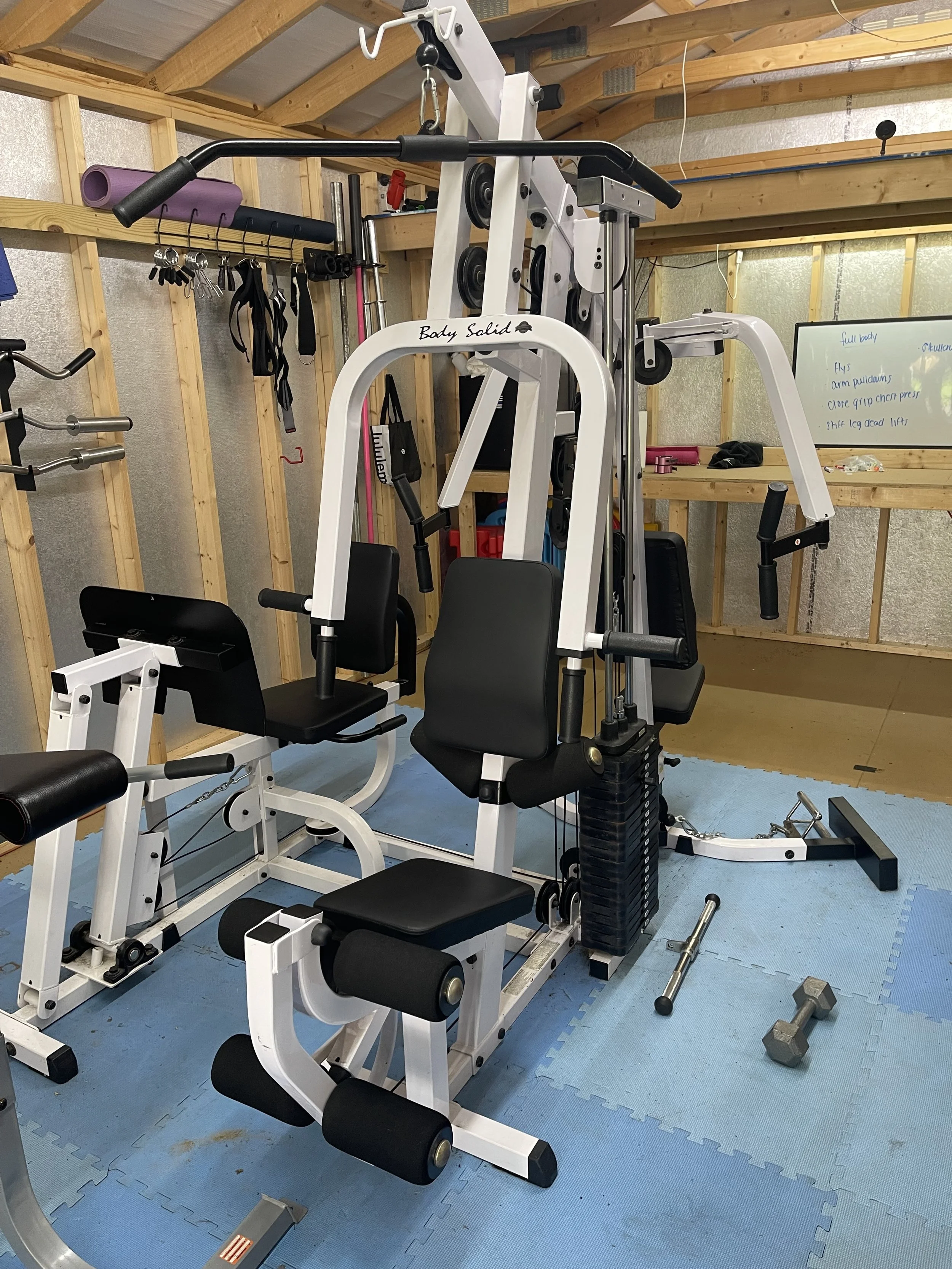 Home gym with various workout equipment including a seated leg curl machine, weights, and a whiteboard on a shelving unit.