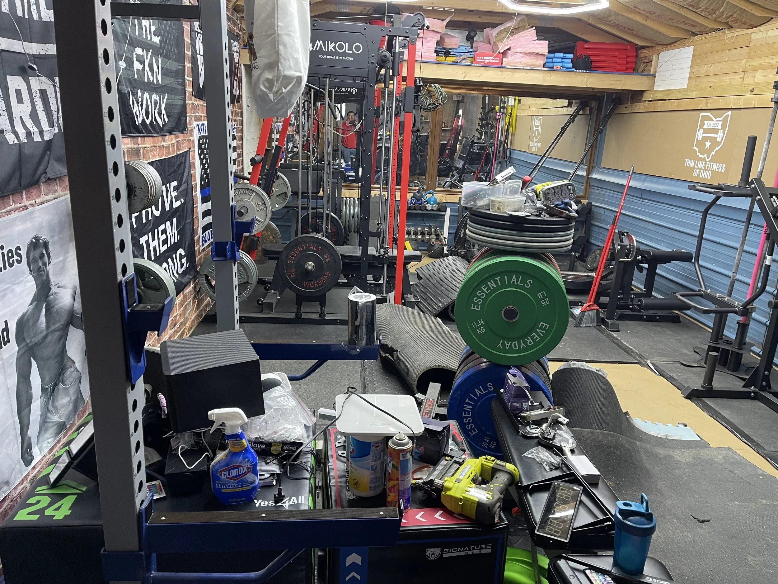 A cluttered gym workspace with various weights, equipment, and tools for fitness and maintenance activities, including weights, lifting racks, and cleaning supplies.