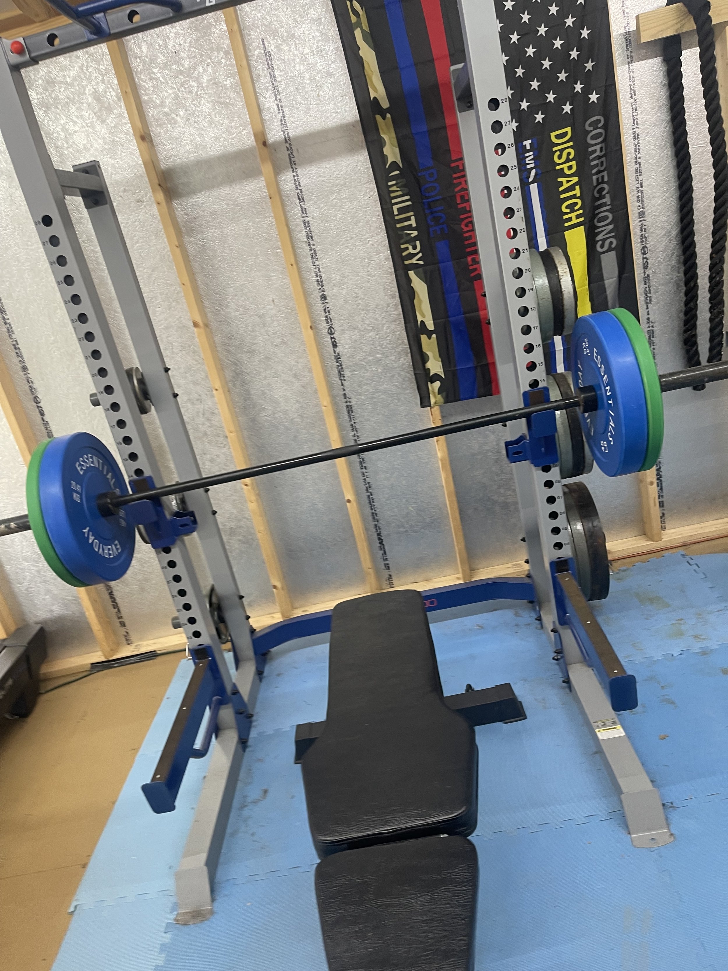A weightlifting bench with a barbell loaded with blue and green weights on a squat rack, with resistance bands hanging on the wall and a padded bench in front.