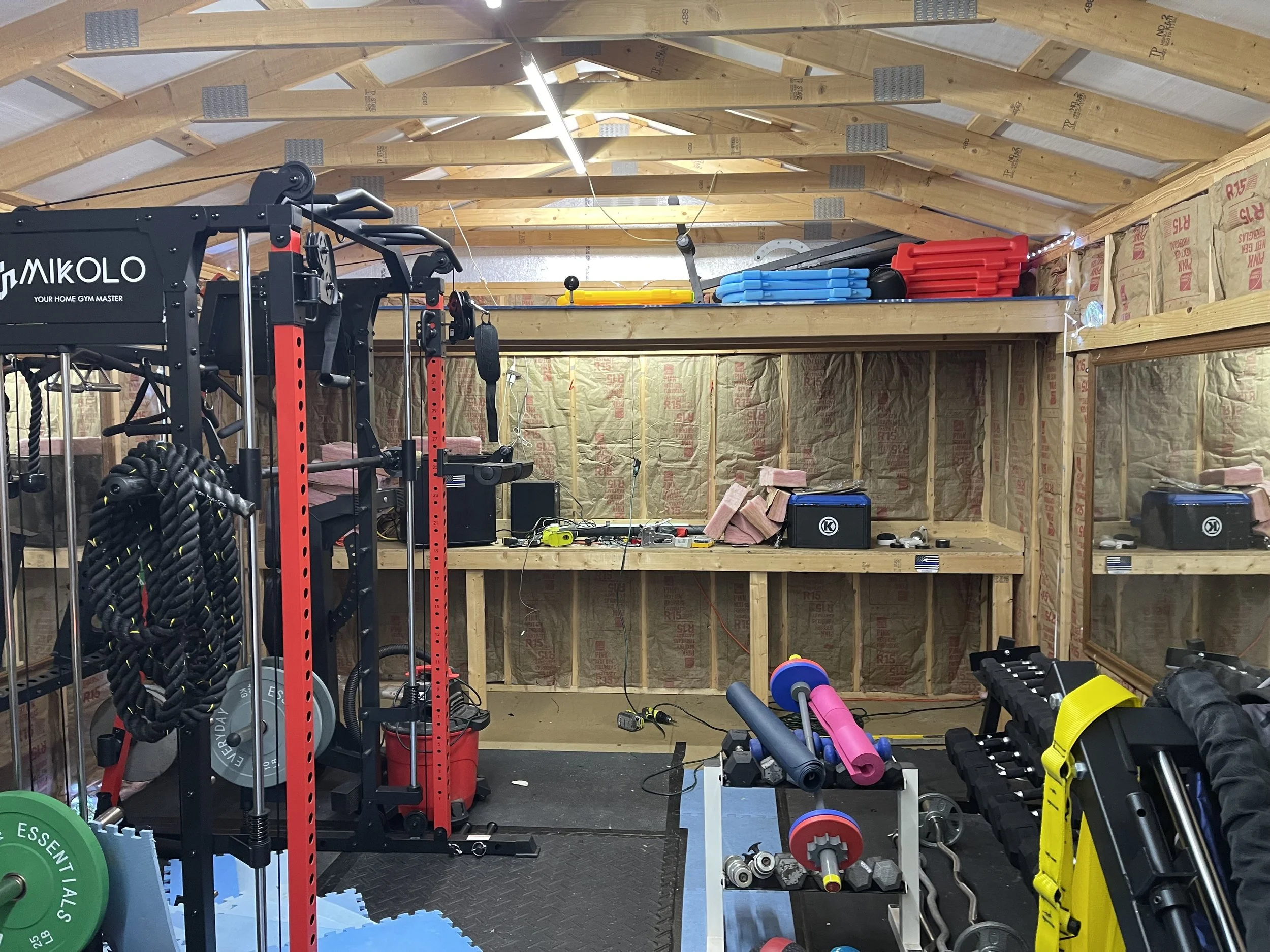 Home gym in a partially finished wooden building with workout equipment, weights, exercise mats, and shelves with fitness accessories.
