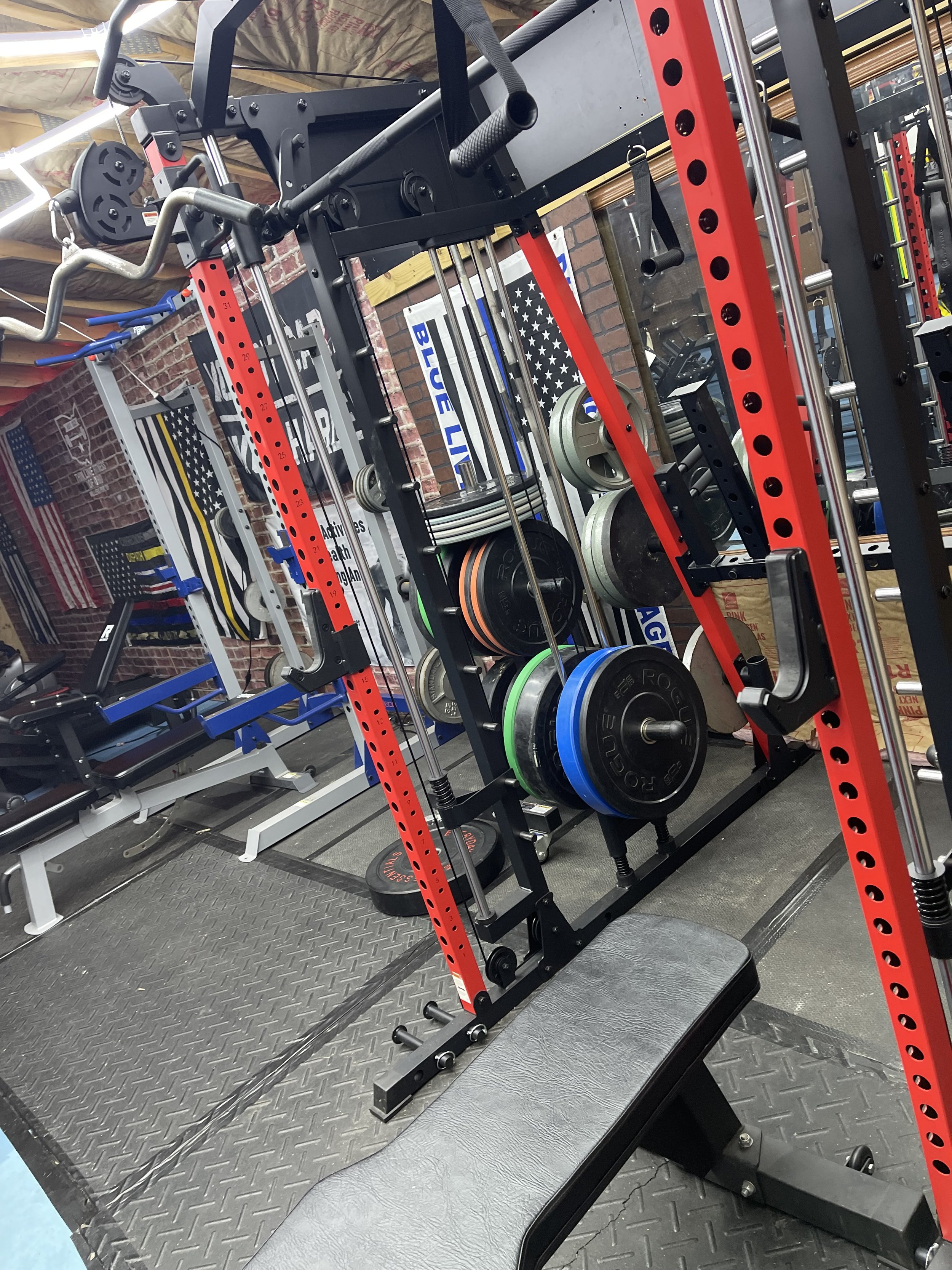 A gym squat rack with weight plates, barbells, and a bench, surrounded by fitness equipment and American flag banners.